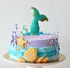 Single-tier round mermaid cake with teal tail topper, lavender icing, aqua wave ruffles, seashells, starfish and bubbles