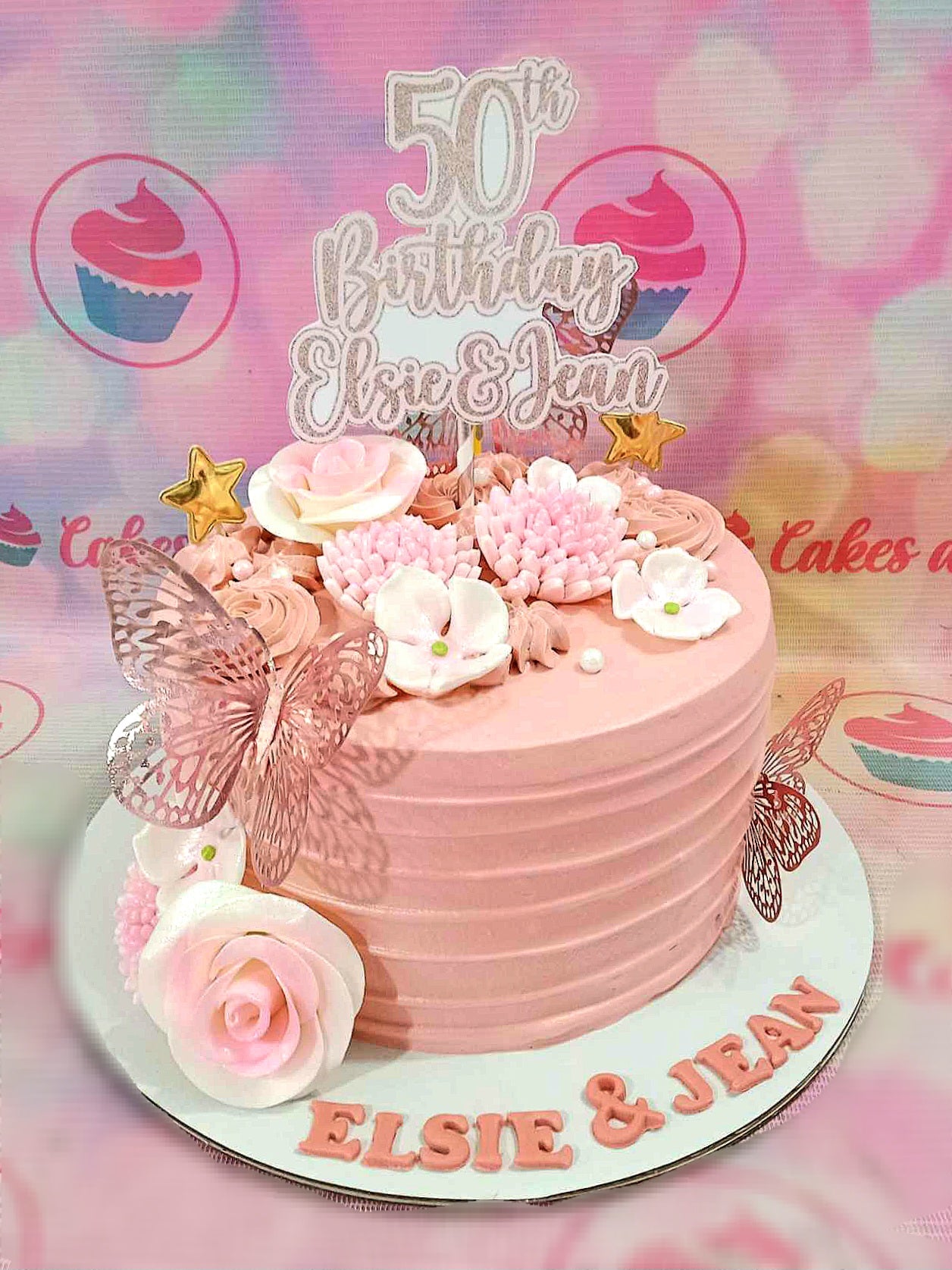 This elegant Butterflies Cake features a stunning pink and rose gold design adorned with delicate sugar flowers and roses. Topped with a 50th birthday printout topper, it sparkles with gold stars and intricate butterflies, making it perfect for anniversaries and celebrations.