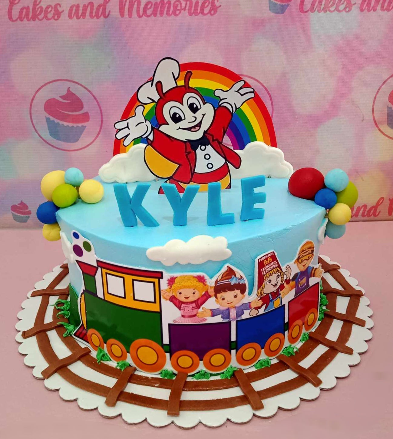 This custom Jolibee Cake features a colorful design with blue, white, red, yellow, and green, showcasing a Jollibee and friends train amidst balloon and cloud accents. Perfect for a baby boy's 1st birthday, the one-tier cake is adorned with playful printout toppers, capturing the joy of the celebration.