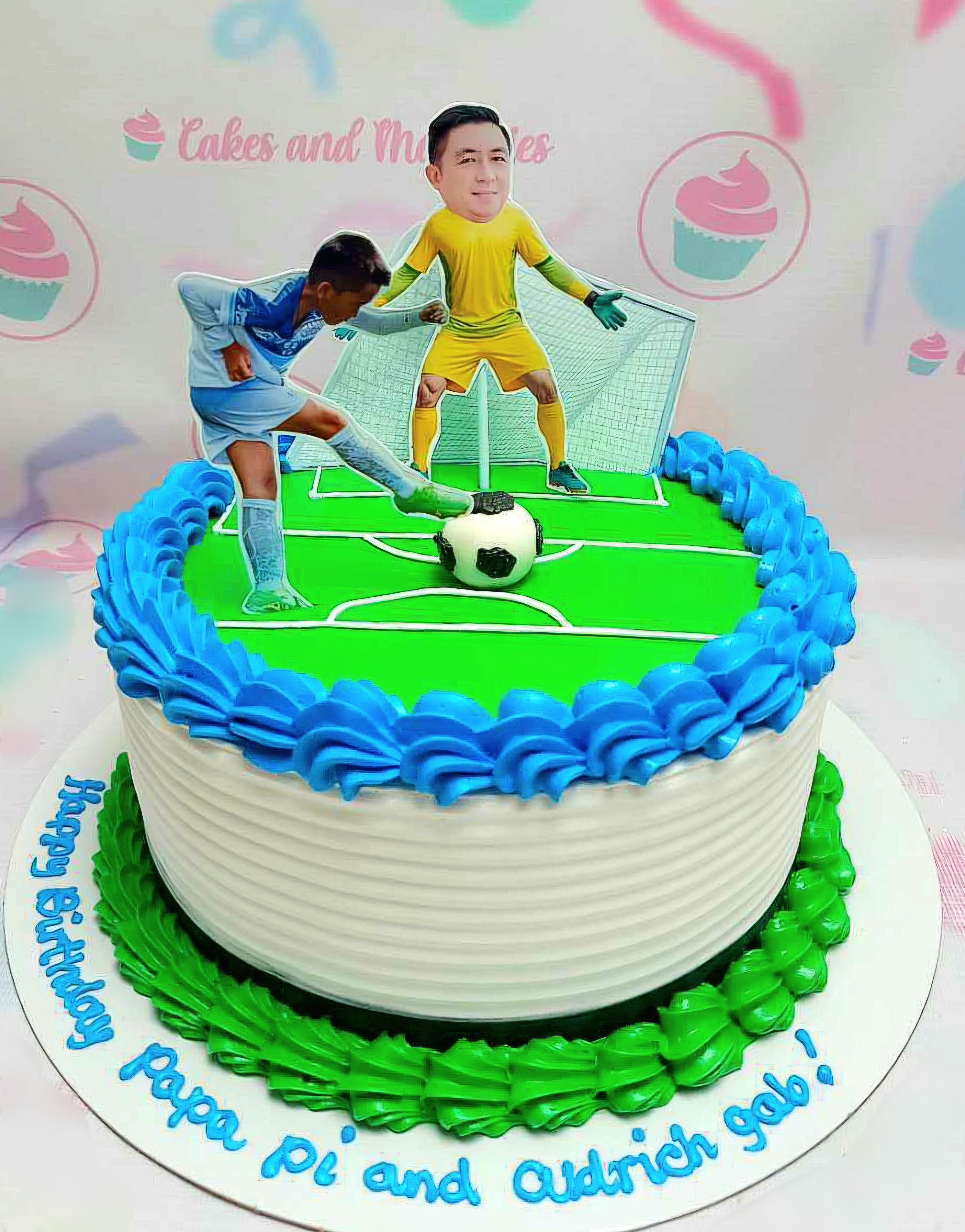 This soccer-themed cake features a white base with green icing, showcasing a detailed soccer field, a soccer ball, and football players including a goalie. Accented with blue details and printout toppers, it's the perfect centerpiece for any soccer celebration.