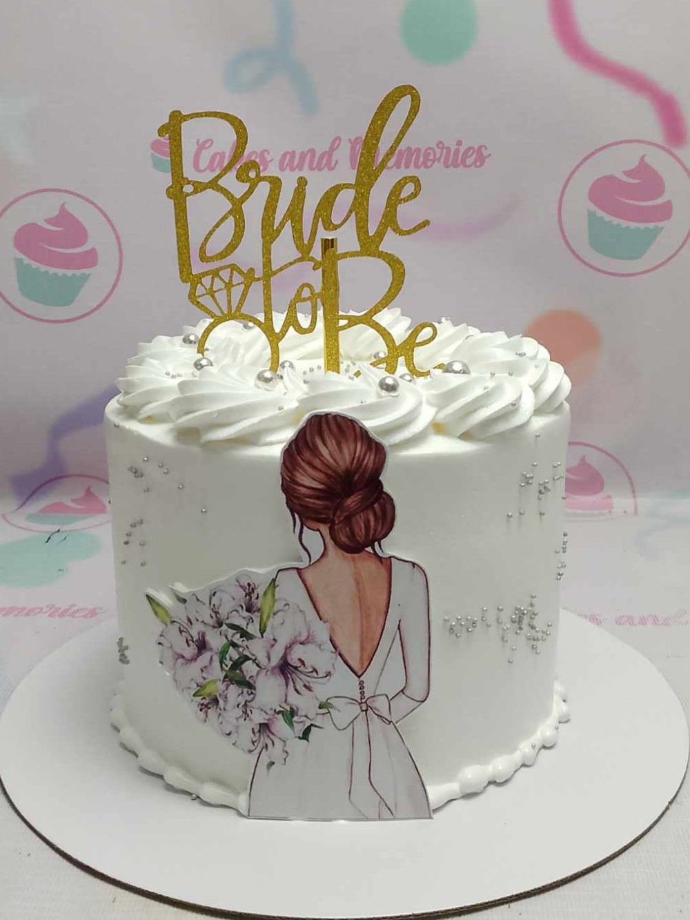 This stunning white bride cake features a gold glitter topper and elegant silver beads, perfect for a bridal shower or bachelorette party. The charming printout toppers add a personal touch, making it an exquisite centerpiece for the bride-to-be's celebration.