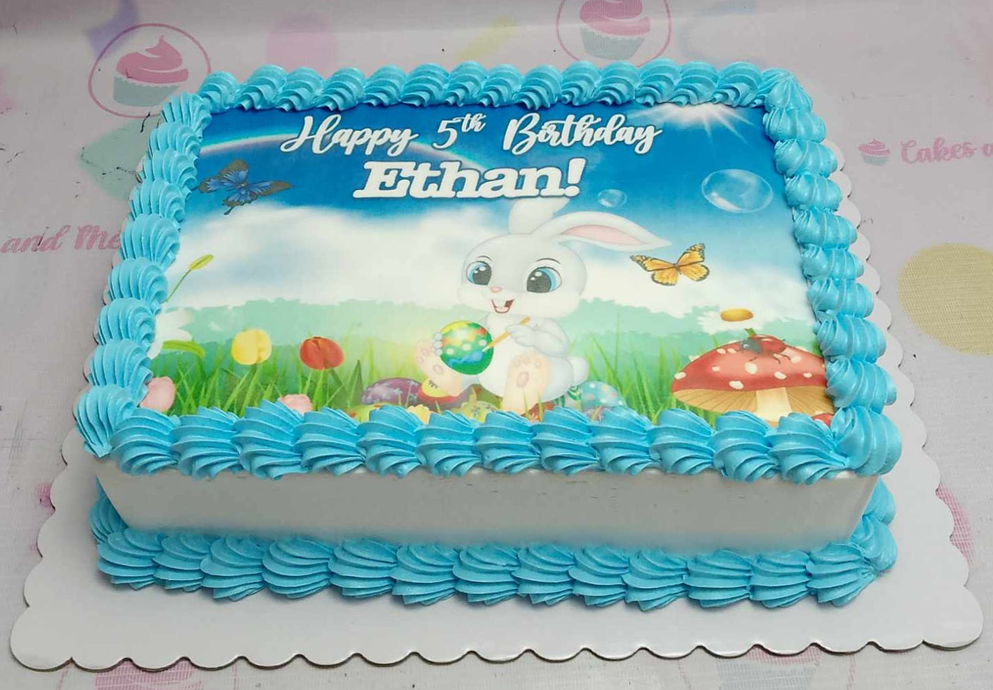 This Bunny Cake features a charming rectangular design adorned with an edible photo print highlighting a playful rabbit and colorful Easter eggs, perfect for a 5th birthday celebration. The soft blue backdrop adds a delightful touch to the festive theme.