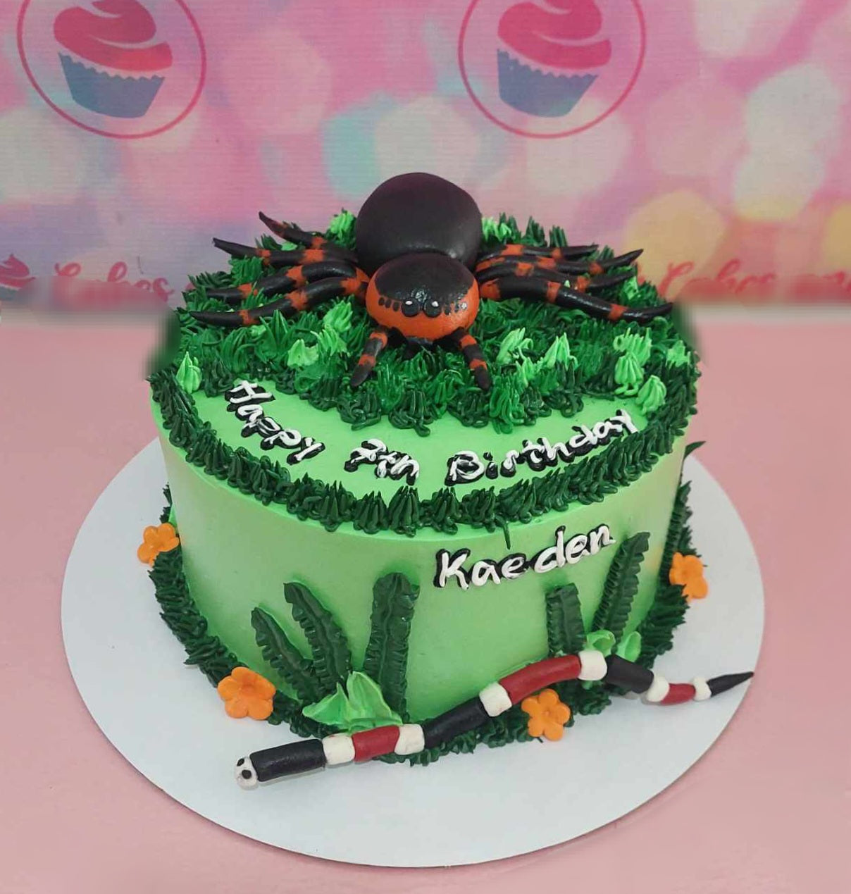 This whimsical Spider Cake features a playful green design with a cute spider and snake atop, perfect for a 7th birthday celebration. Adorned with lush grass and colorful frosting, this one-layer, one-tier cake is an eye-catching centerpiece.