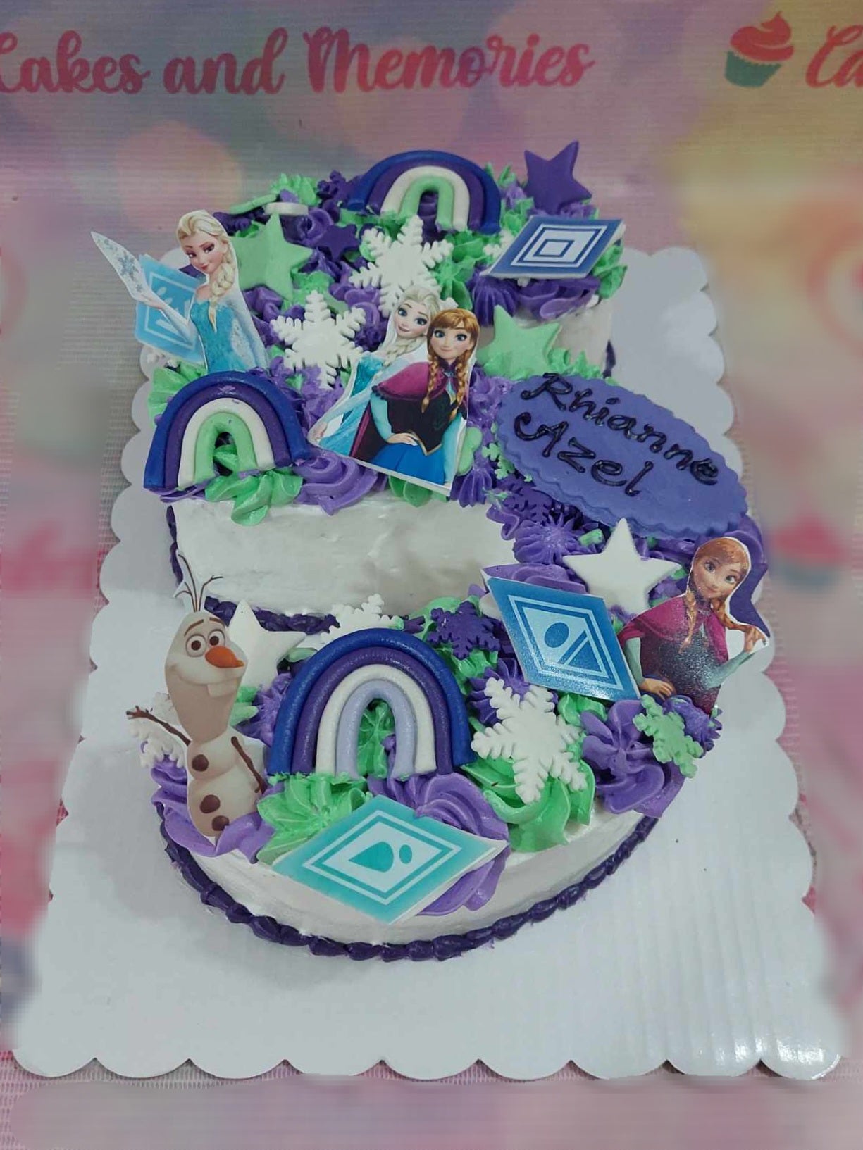 This vibrant rectangular Frozen-themed cake features a whimsical blend of purple, green, and blue shades, adorned with charming stars and snowflakes. Decorated with playful printout toppers of Elsa, Anna, and Olaf, it's an enchanting centerpiece for any 5th birthday celebration.