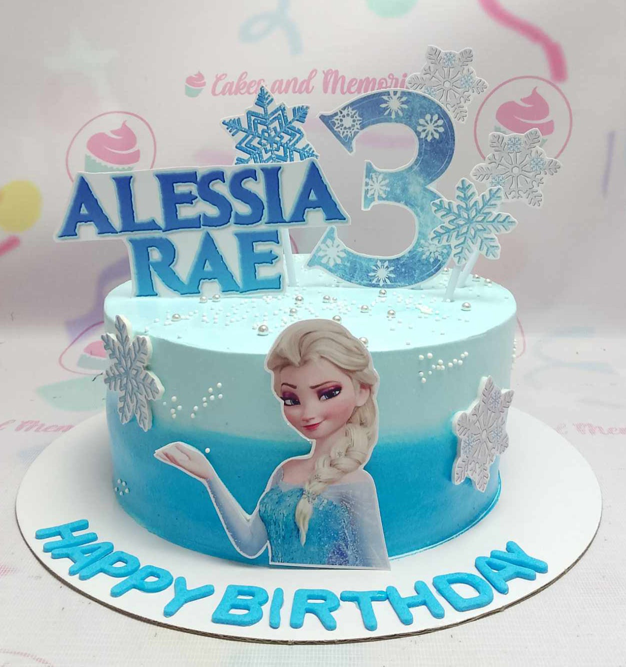 This custom Frozen cake features a round design adorned in blue and white frosting, showcasing snowflakes and printout toppers of Elsa and Anna. Celebrating "happy birthday" for Alessia Rae with the number "3," this delightful one-tier cake is perfect for any children's birthday party.