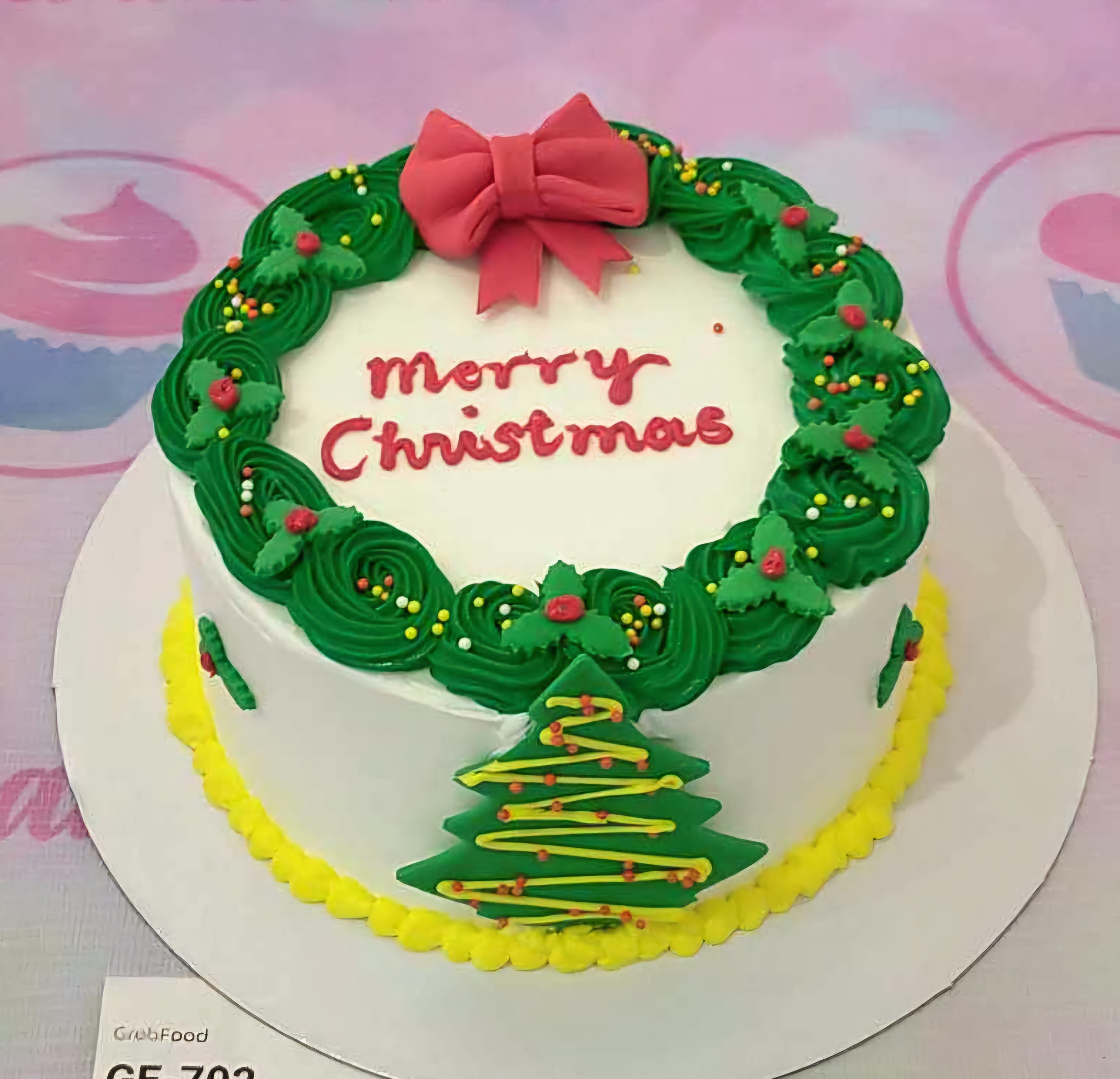 This festive Christmas cake features a sleek white design adorned with a wreath and a red bow, complemented by sprinkles and a cheerful yellow border. Perfect for any Christmas party, it is a single-layer, single-tier cake that captures the holiday spirit.