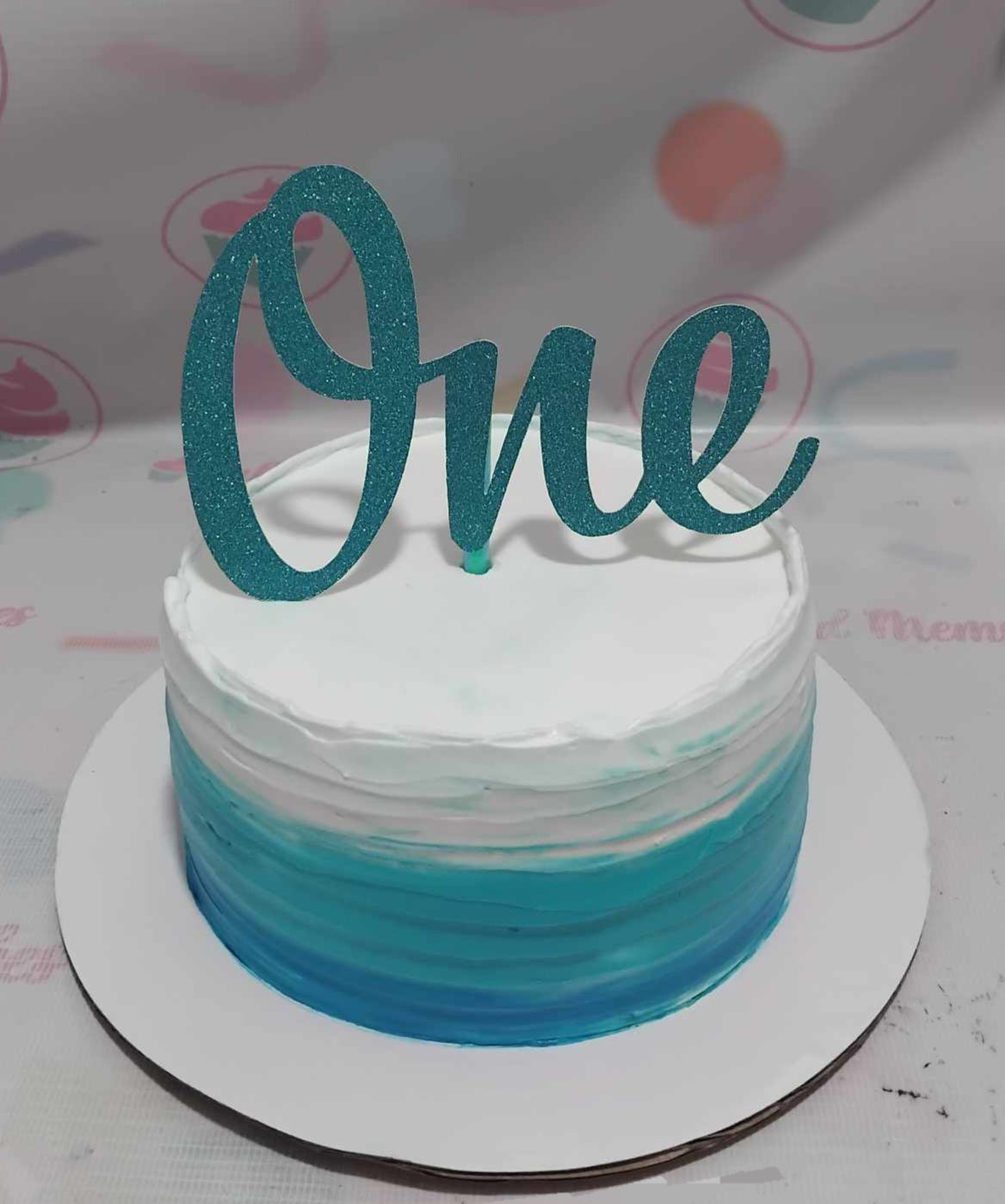 This stunning one-tier cake features a beautiful blend of blue and teal hues, adorned with a sparkling glitter topper celebrating a first birthday. Perfect for a cake smash, this one-layer design is both eye-catching and festive.