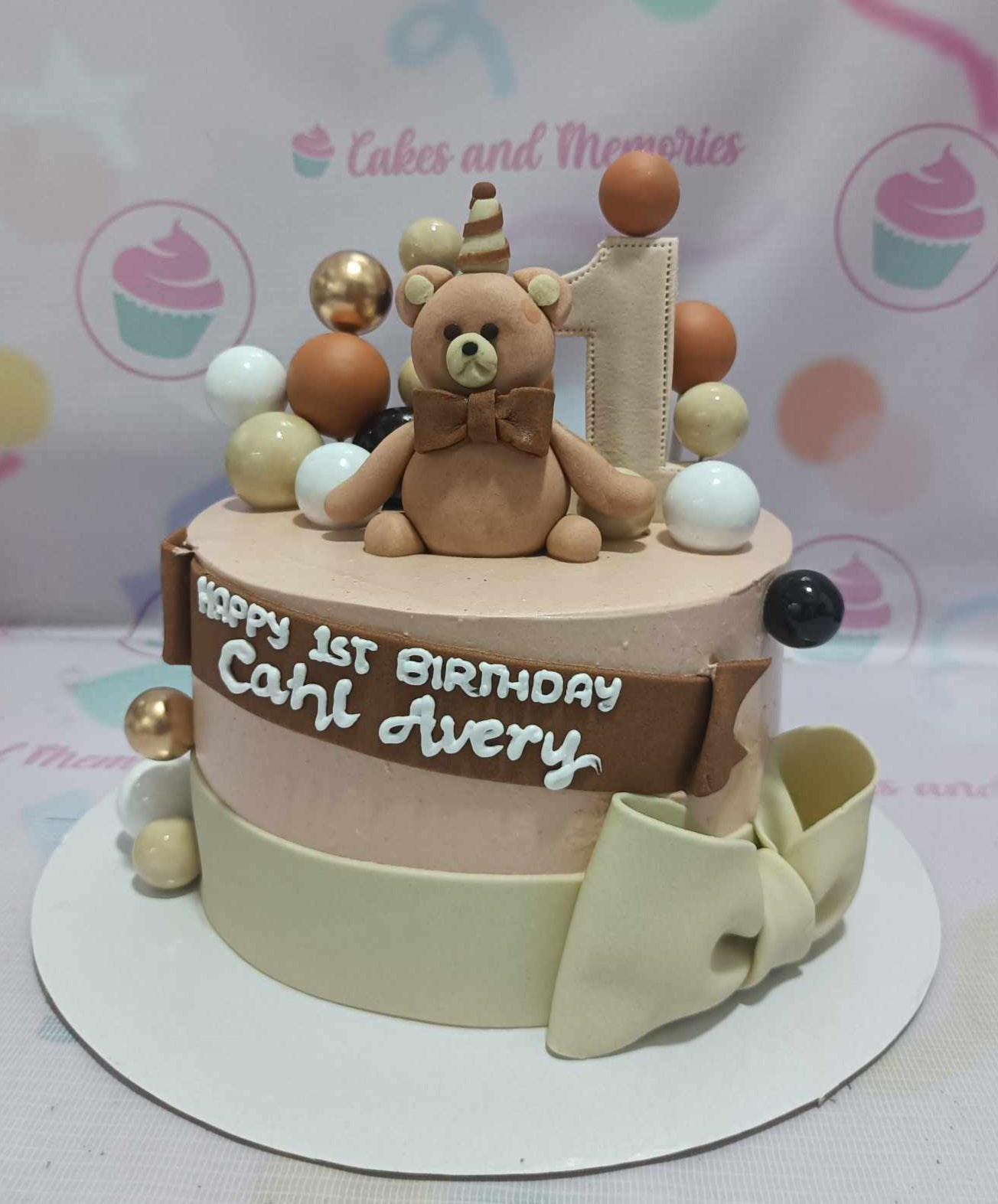 A charming 1st birthday Bears Cake featuring a cute teddy bear design in rich brown and gold hues, adorned with playful ball decorations and ribbon accents. The one-tier cake is topped with delightful printout toppers of wild animals, perfect for a special celebration.