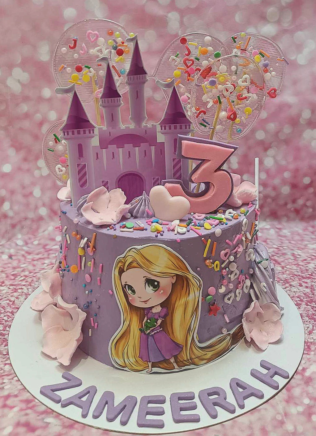 This enchanting Rapunzel Cake features shades of lavender and purple, adorned with lollipops, sprinkles, and pink flowers, perfect for Zameerah's 3rd birthday celebration. The single-tier design showcases charming printout toppers and a castle, capturing the spirit of the beloved fairytale.