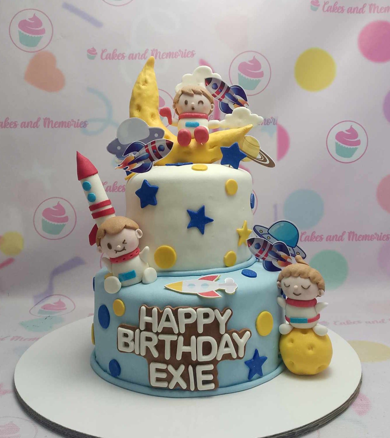 This stunning Space Cake features two layers adorned with blue and white fondant, embellished with yellow stars, planets, and an astronaut. Perfect for Exie's 1st birthday, it showcases a charming spaceship and playful aliens against a celestial backdrop.