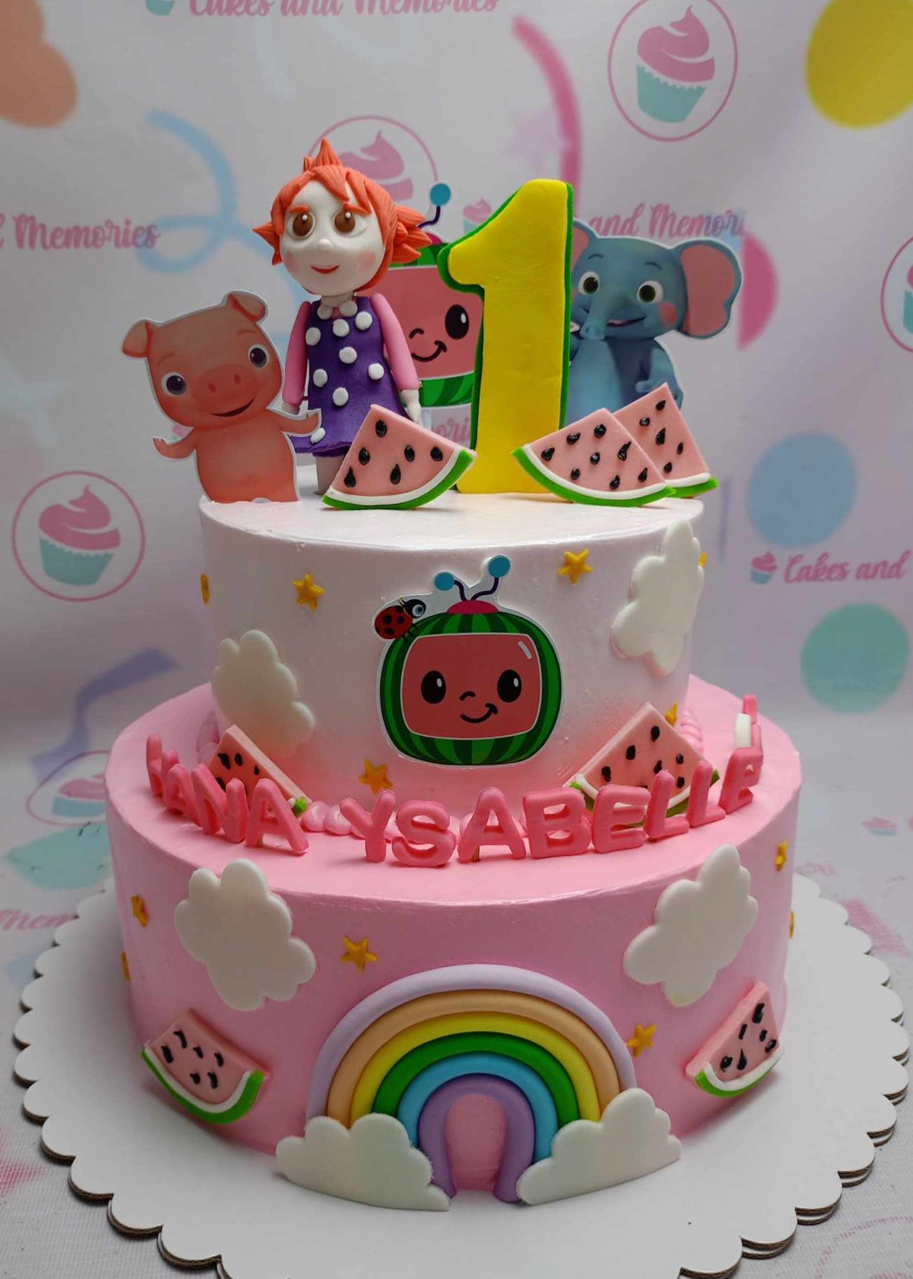 This custom Cocomelon cake features two pink tiers adorned with clouds, an edible rainbow, and playful printout toppers of a pig and elephant, perfect for a baby girl's 1st birthday celebration. The design includes a fun watermelon theme and a prominent "one" to mark this special milestone.