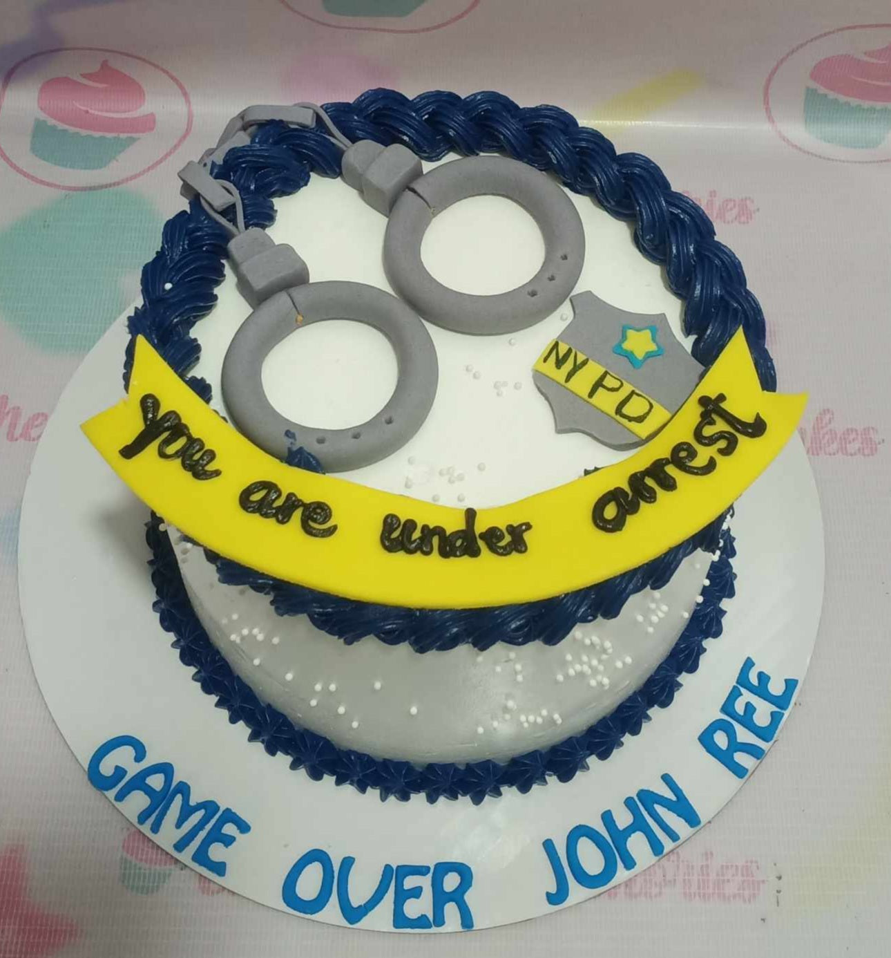 This custom decorated Police Cake features a striking white and blue design, adorned with playful handcuffs and a "Game Over" message, perfect for a bachelor's or stag party. The one-tier creation captures a mischievous spirit with icons like guns and a "Under Arrest" theme, making it an ideal centerpiece for any fun celebration.