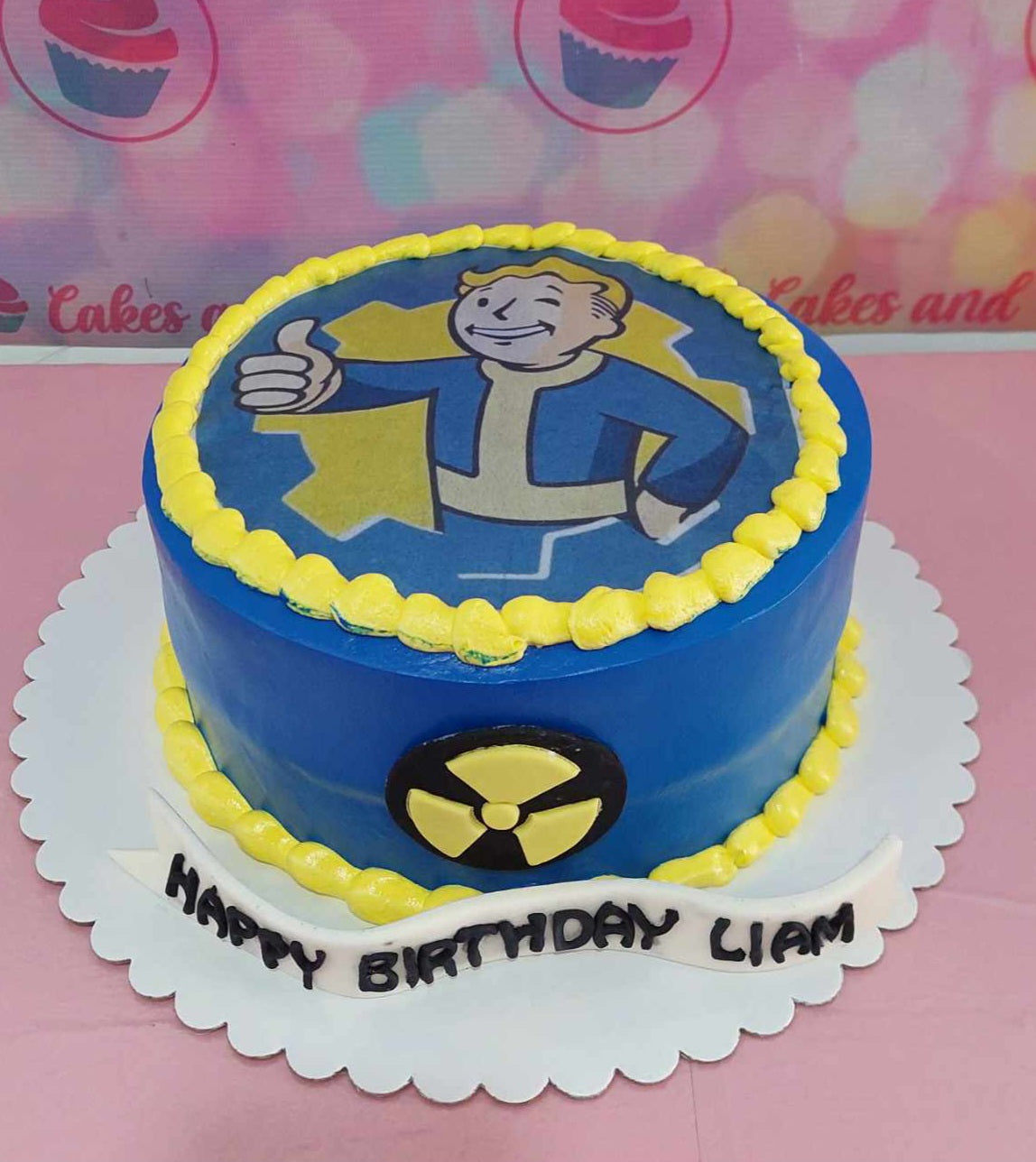 This single-tier Video Games Cake features a striking design in blue and yellow, showcasing an edible image of the Fallout vault boy, complete with a radioactive symbol. Perfect for celebrating "Happy Birthday Liam," this cake captures the essence of gaming with themed printout toppers and references to popular titles like PUBG.