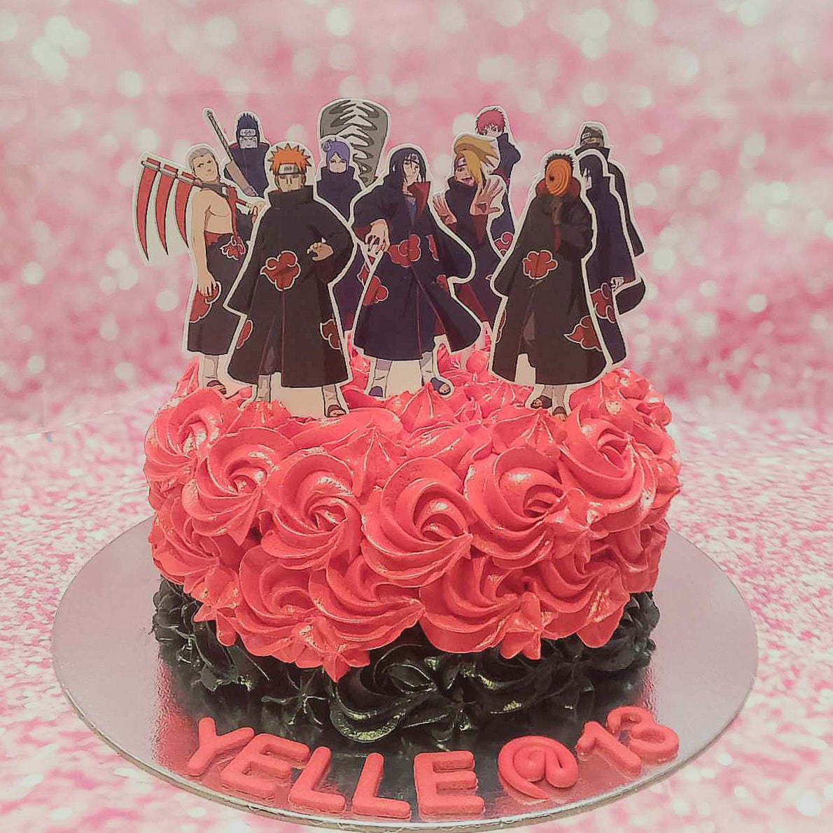 A striking Naruto-themed cake adorned with red and black rosettes, featuring Akatsuki details and printed Naruto character toppers for Yelle's 13th birthday. This one-tier masterpiece captures the essence of anime in its bold design.
