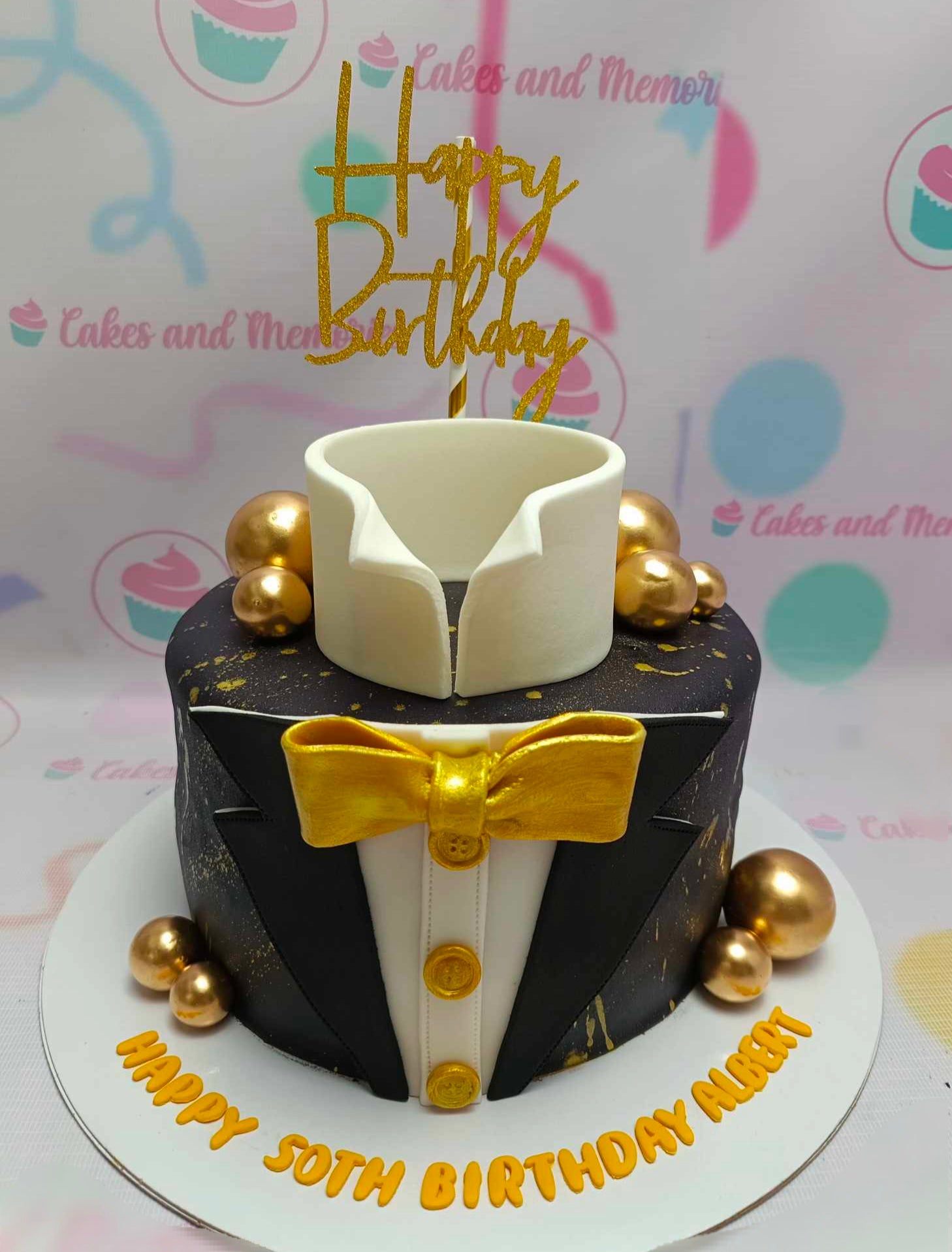 This elegant suit cake features a black tuxedo design complete with a bowtie, gold buttons, and decorative gold spheres, perfect for celebrating Albert's 50th birthday. The single-tier fondant creation showcases sophistication and style.