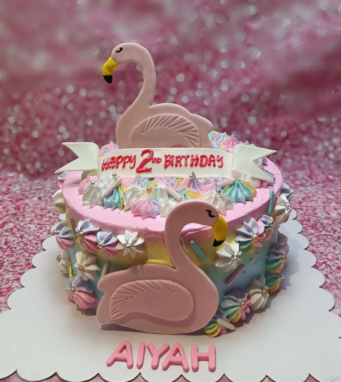 A stunning pink Flamingo Cake designed for Aiyah's 2nd birthday, featuring adorable flamingos, delicate meringue, colorful sprinkles, and elegant flowers adorned with pearls. This one-tier masterpiece captures a pastel rainbow theme that perfectly complements the celebration.