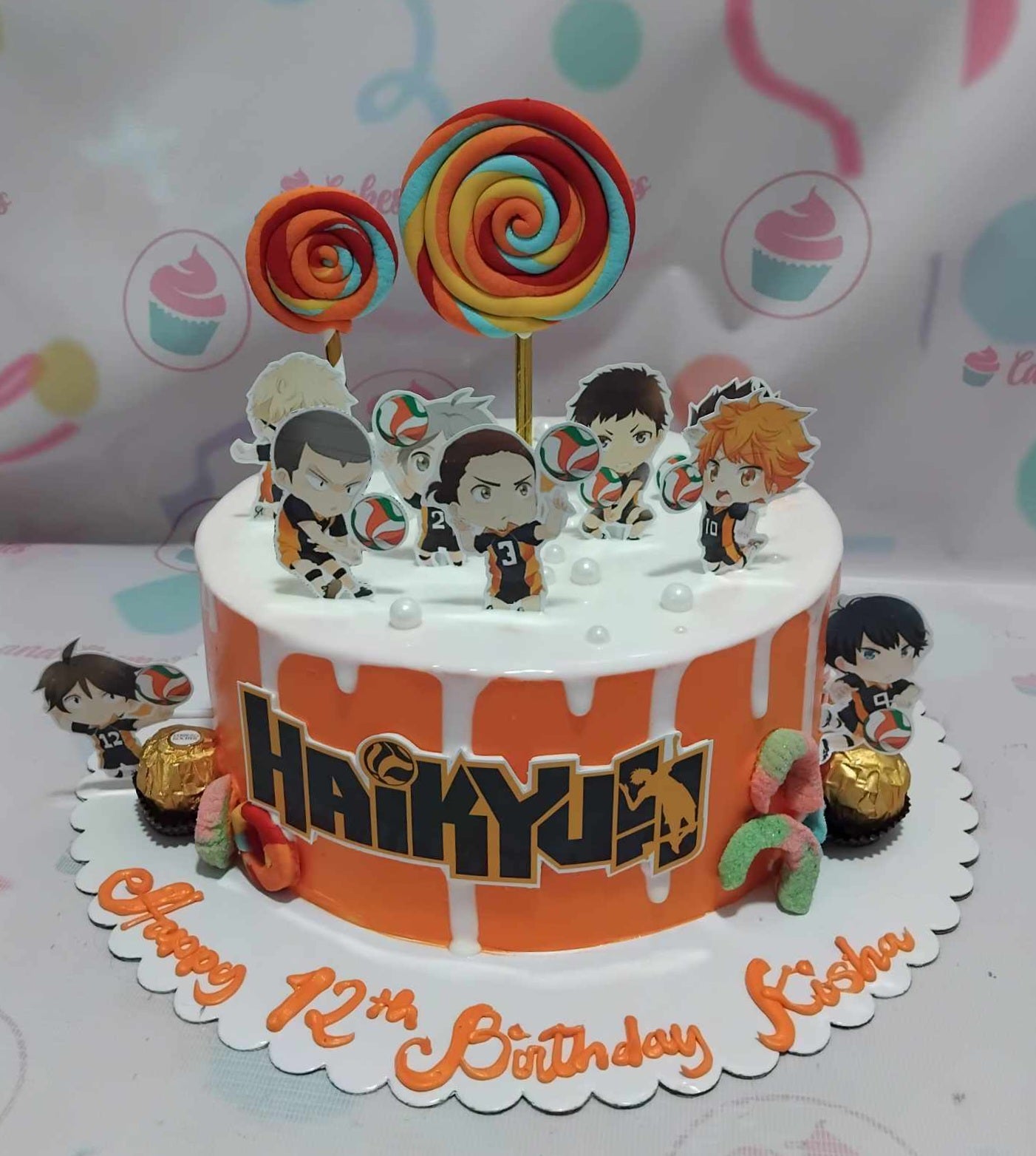 This vibrant one-tier anime cake features a striking orange and white drip design, adorned with colorful lollipops and sports-themed details, including a volleyball and chibi characters from Haikyu. Printout toppers complete the playful look, capturing the essence of Japanese anime and manga.