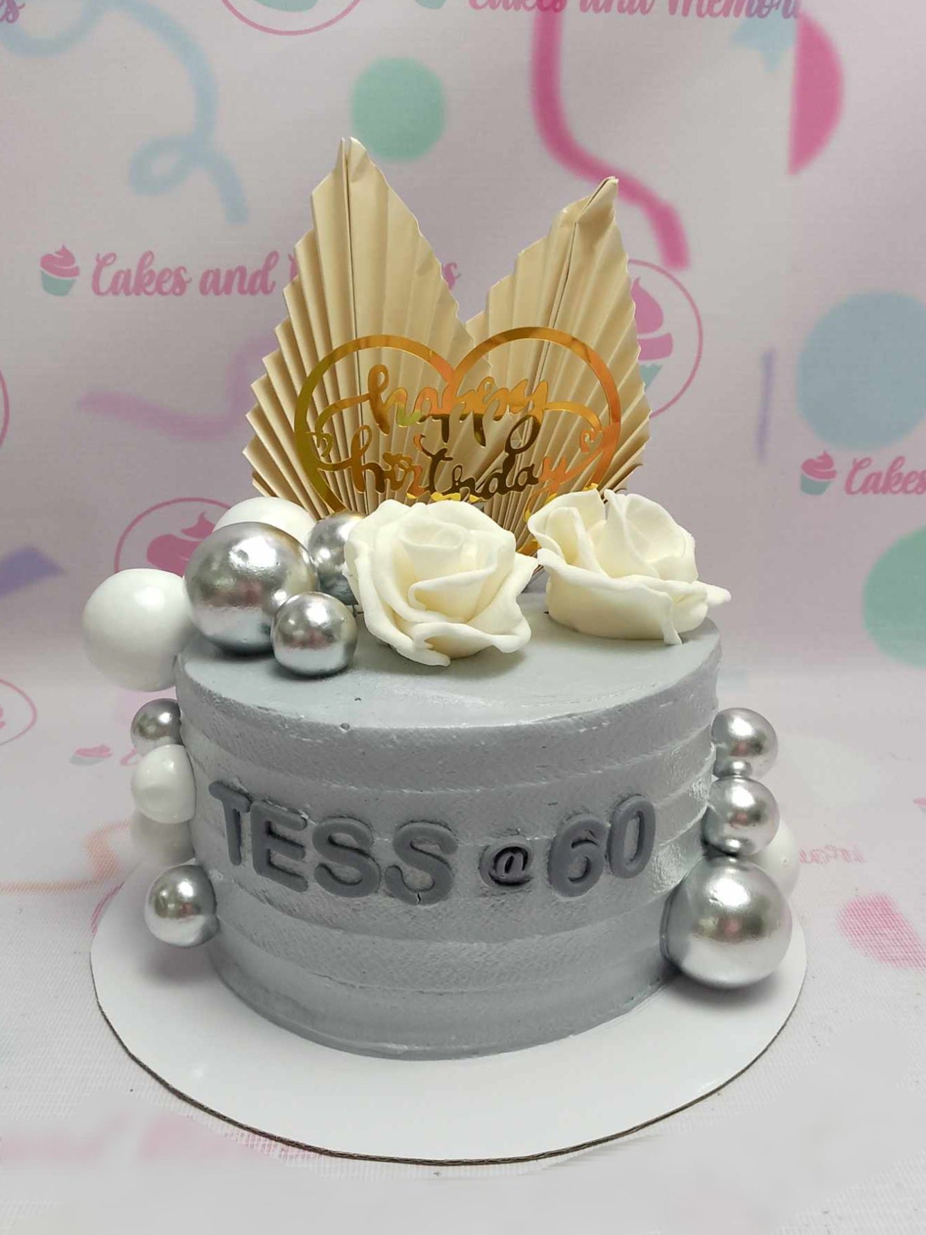 A stunning 1-tier Fifty Cake designed for a 60th birthday celebration, featuring elegant white roses, gray accents, and adorned with silver and white balls. Accented with a golden fan and charming printout toppers, this cake exudes sophistication.