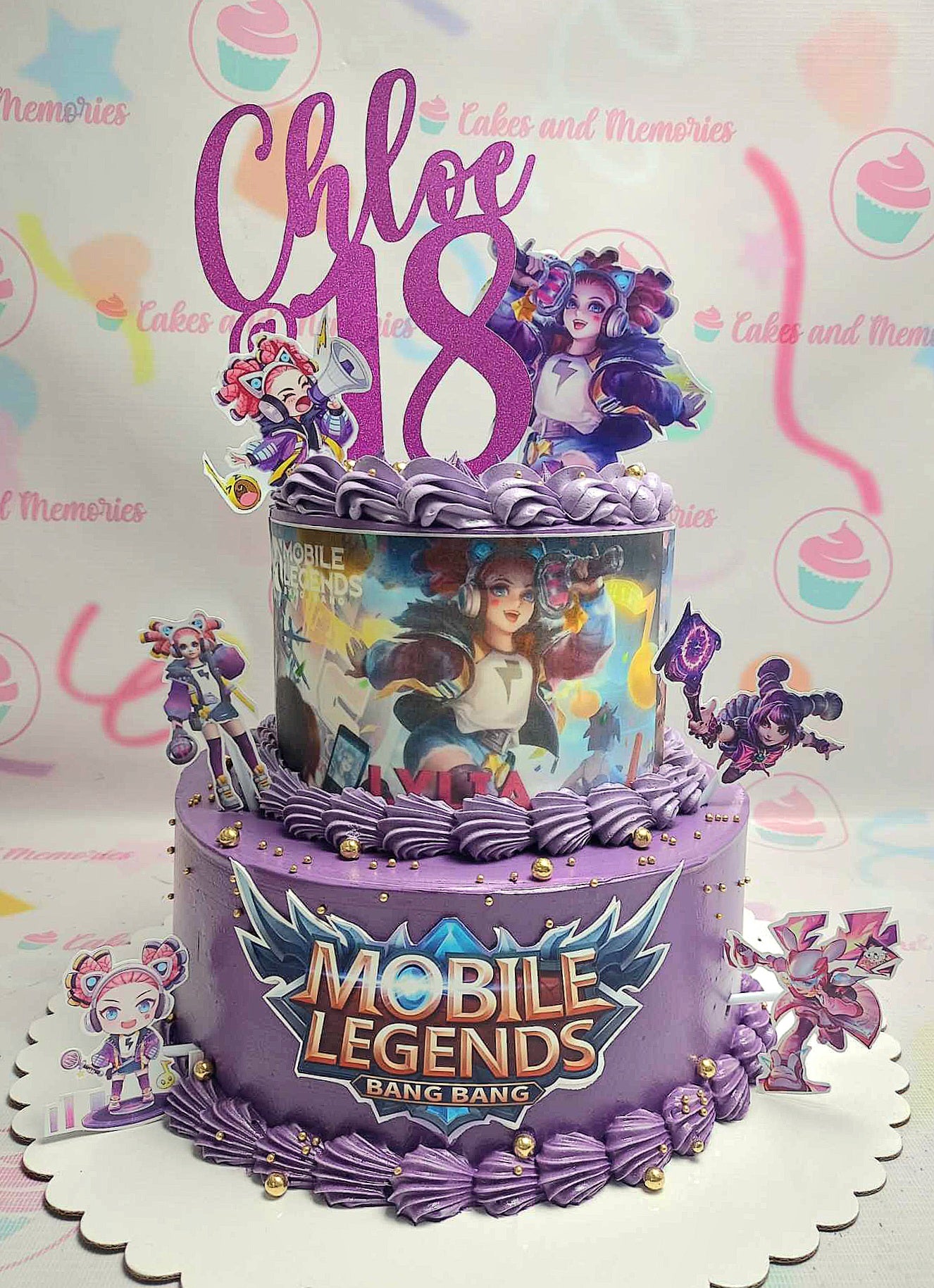 This stunning two-tier Mobile Legends cake features a rich purple fondant base adorned with gold stars and hearts, beautifully showcasing Chloe and Lylia printout characters. Perfect for an 18th birthday celebration, this custom cake combines elegance with a playful theme.