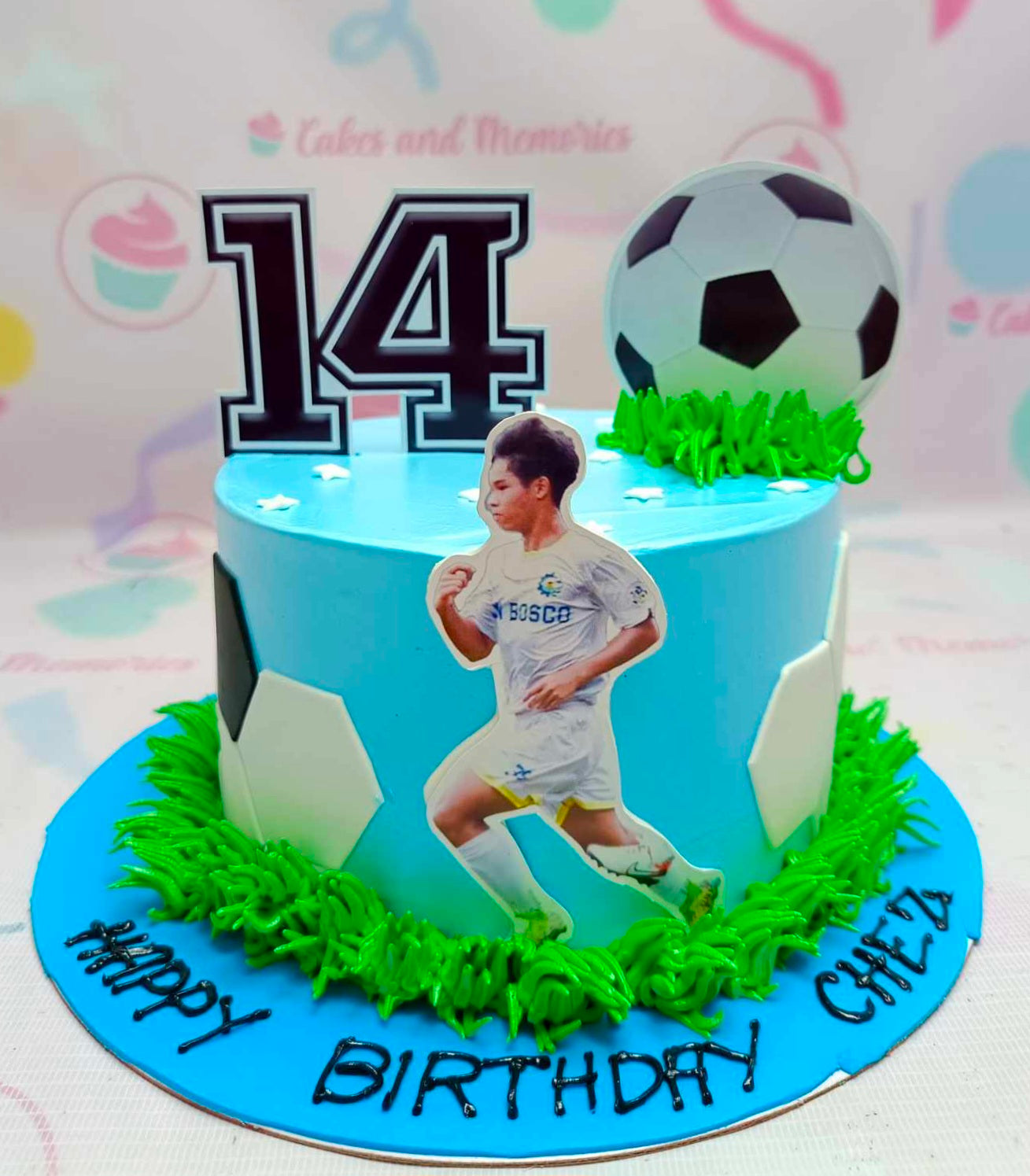 This custom soccer cake features vibrant blue accents with green grass detail, perfect for celebrating a birthday. It's a fun one-tier design adorned with printout toppers, ideal for any sports-loving gentleman or young football fan.