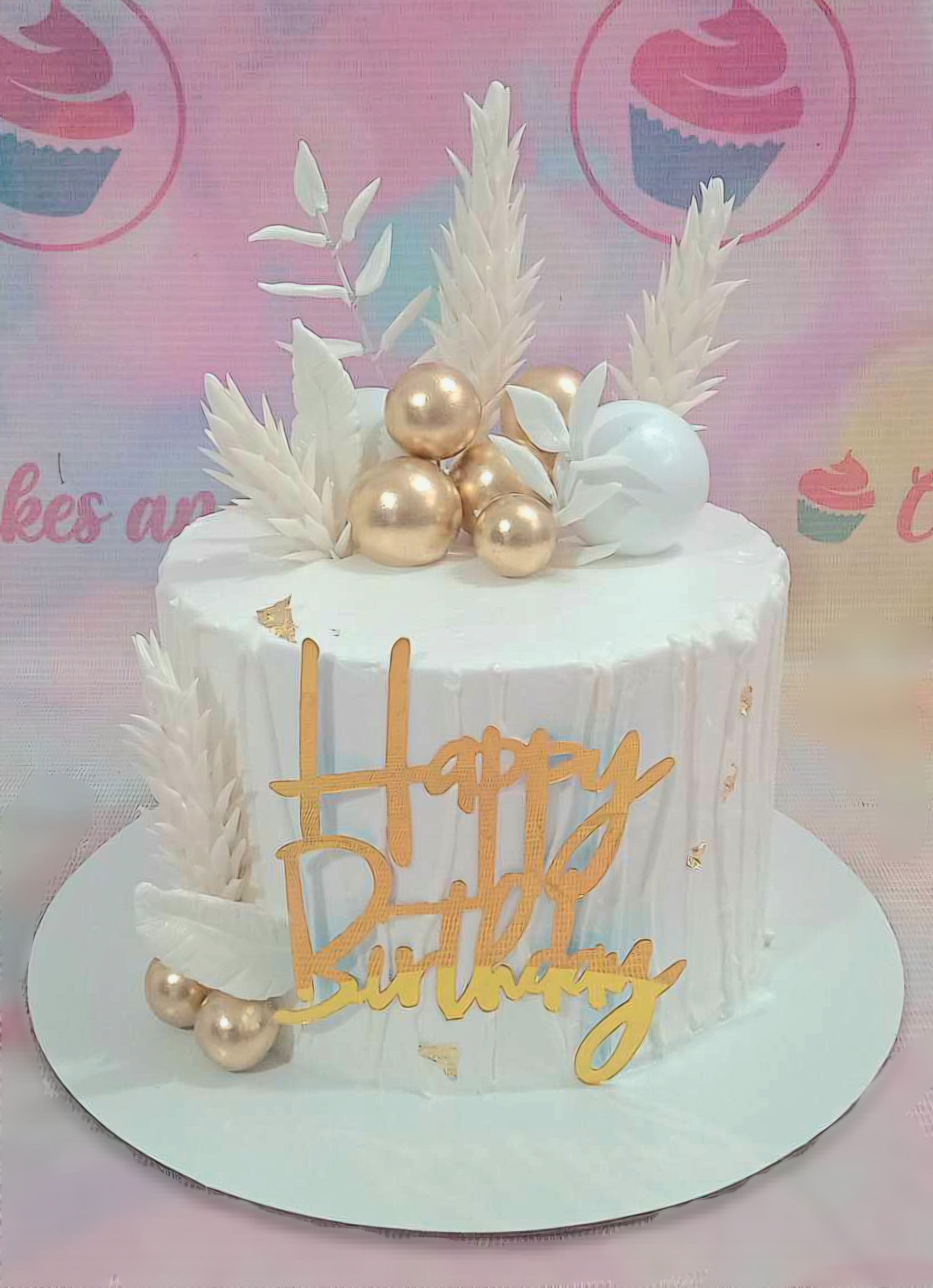 This vibrant Fifty Cake features a sleek white design adorned with gold balls, gold sprinkles, and a happy birthday gold topper, complemented by decorative edible leaves and whimsical feathers. Perfect for celebrating a beloved grandfather or grandmother in a modern and contemporary style.