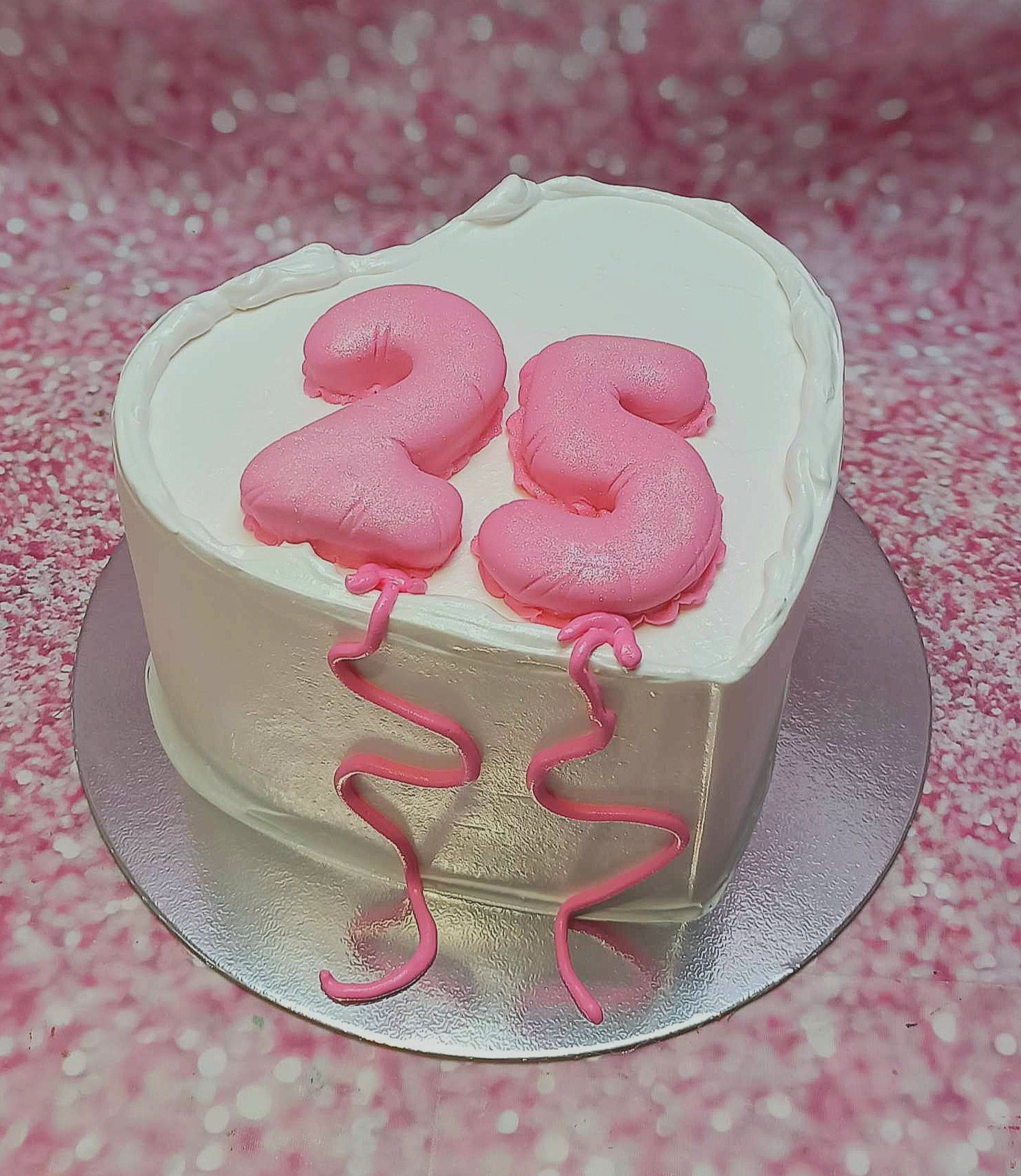 This beautiful heart-shaped Love Cake features smooth white icing adorned with pink ribbons and a cheerful pink balloon. Perfect for a 25th birthday celebration, it proudly displays the number "25" atop the elegant 1-tier design.