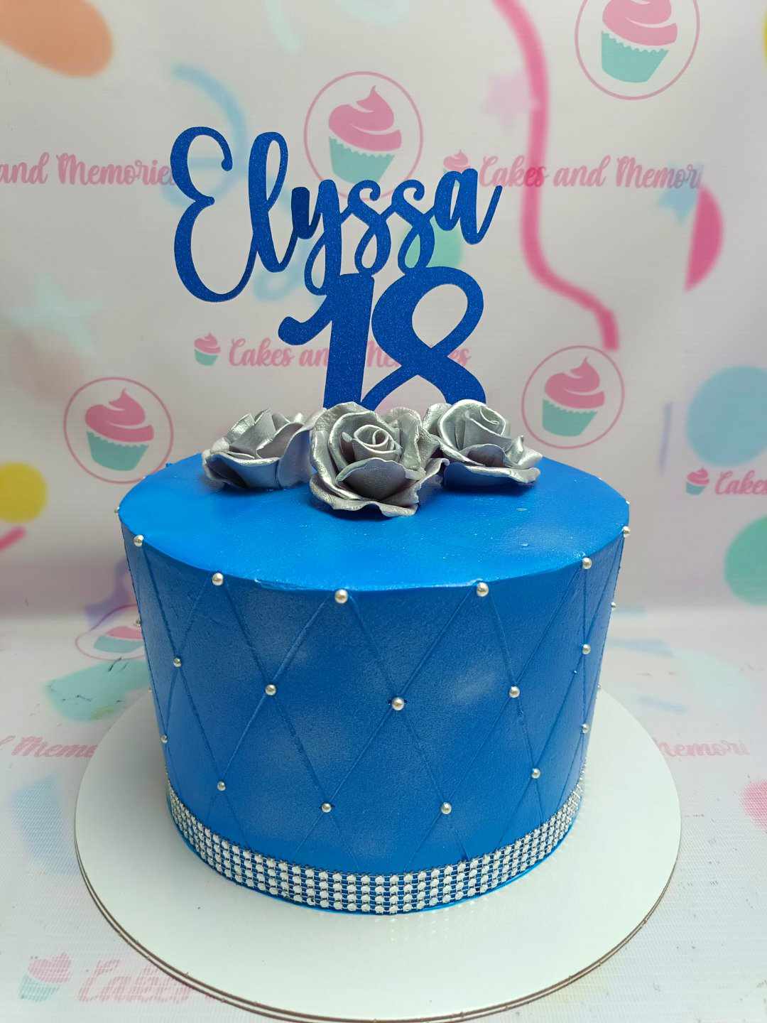 This stunning debut cake features a tall, quilted design in royal blue, adorned with silver roses and an elegant blue glitters cardstock cake topper for Elyssa's 18th birthday. The single tier showcases exquisite detail, perfect for celebrating this milestone occasion.