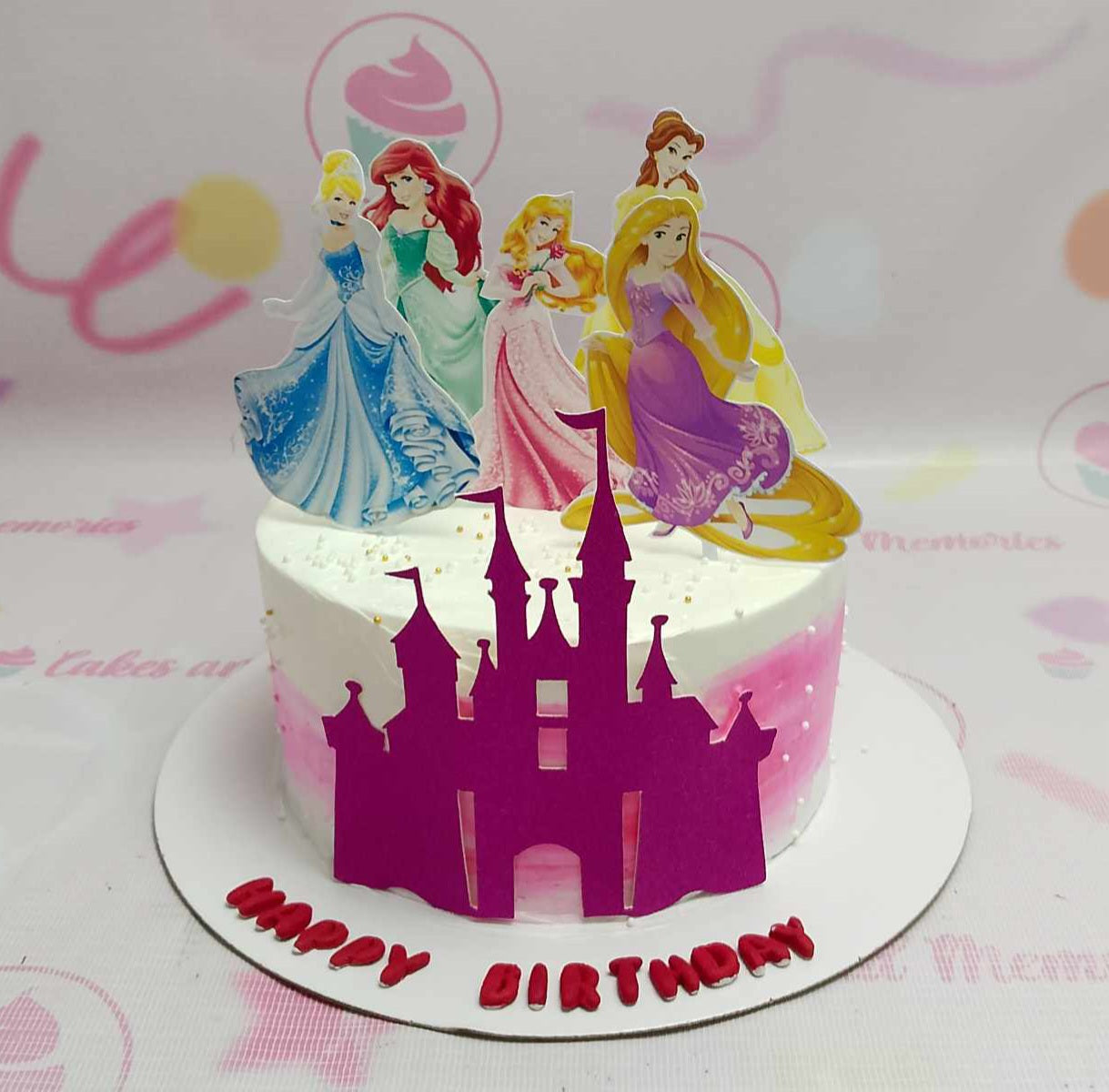 A stunning Princess Cake featuring a pink and white design adorned with gold sprinkles, showcasing printout toppers of Ariel, Cinderella, Belle, Rapunzel, and Aurora. Perfect for a girl's birthday celebration, this one-tier cake brings a magical touch to any event.