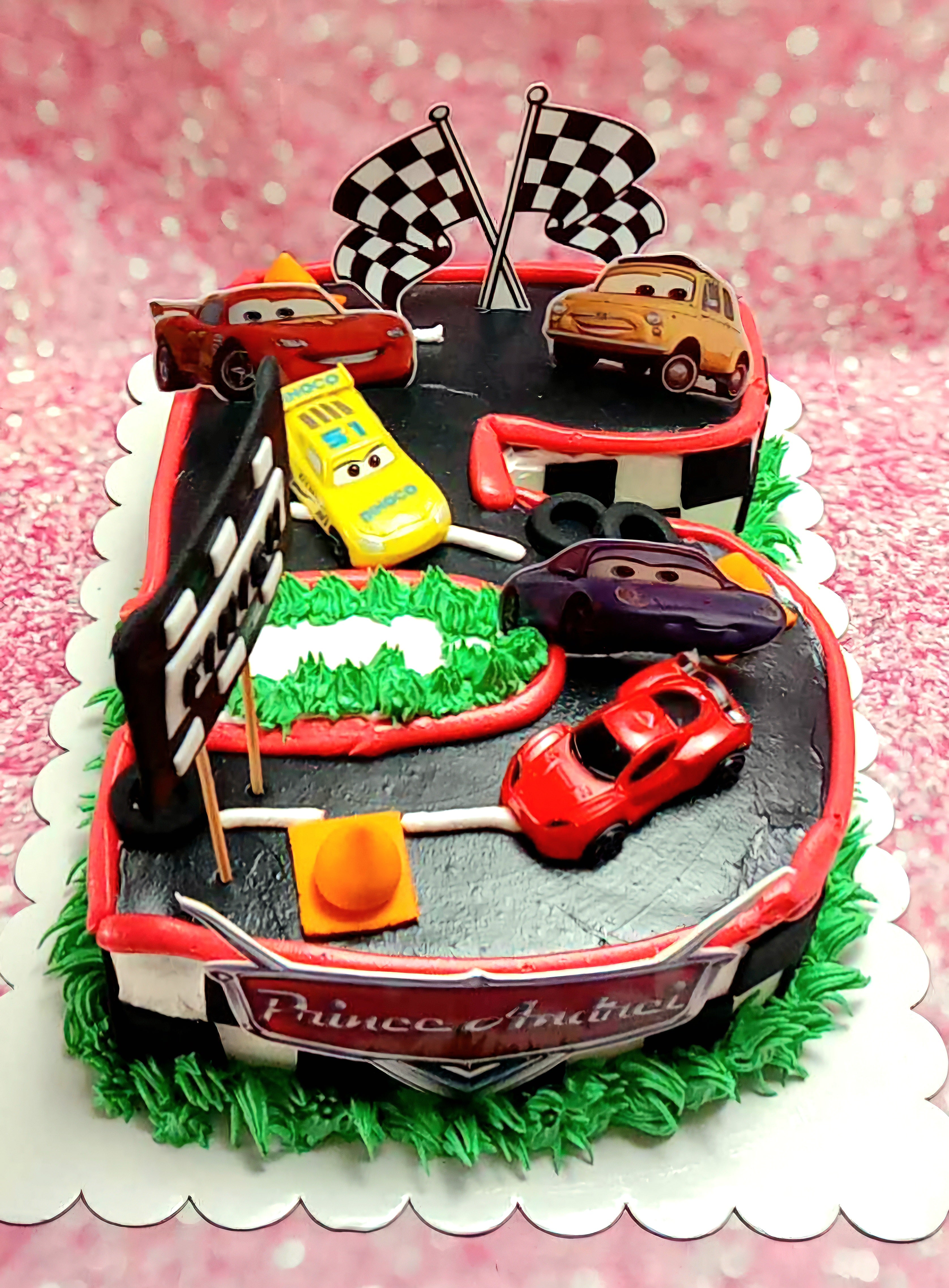 This custom Cars (Pixar) cake features a rectangular design adorned with black, red, and white colors, perfect for a fifth birthday celebration. Showcasing beloved characters like Lightning McQueen, Mater, and Francesco Bernoulli, it’s set against a backdrop of green grass, a race track, checkered flags, cones, and tires, complete with detailed printout toppers.