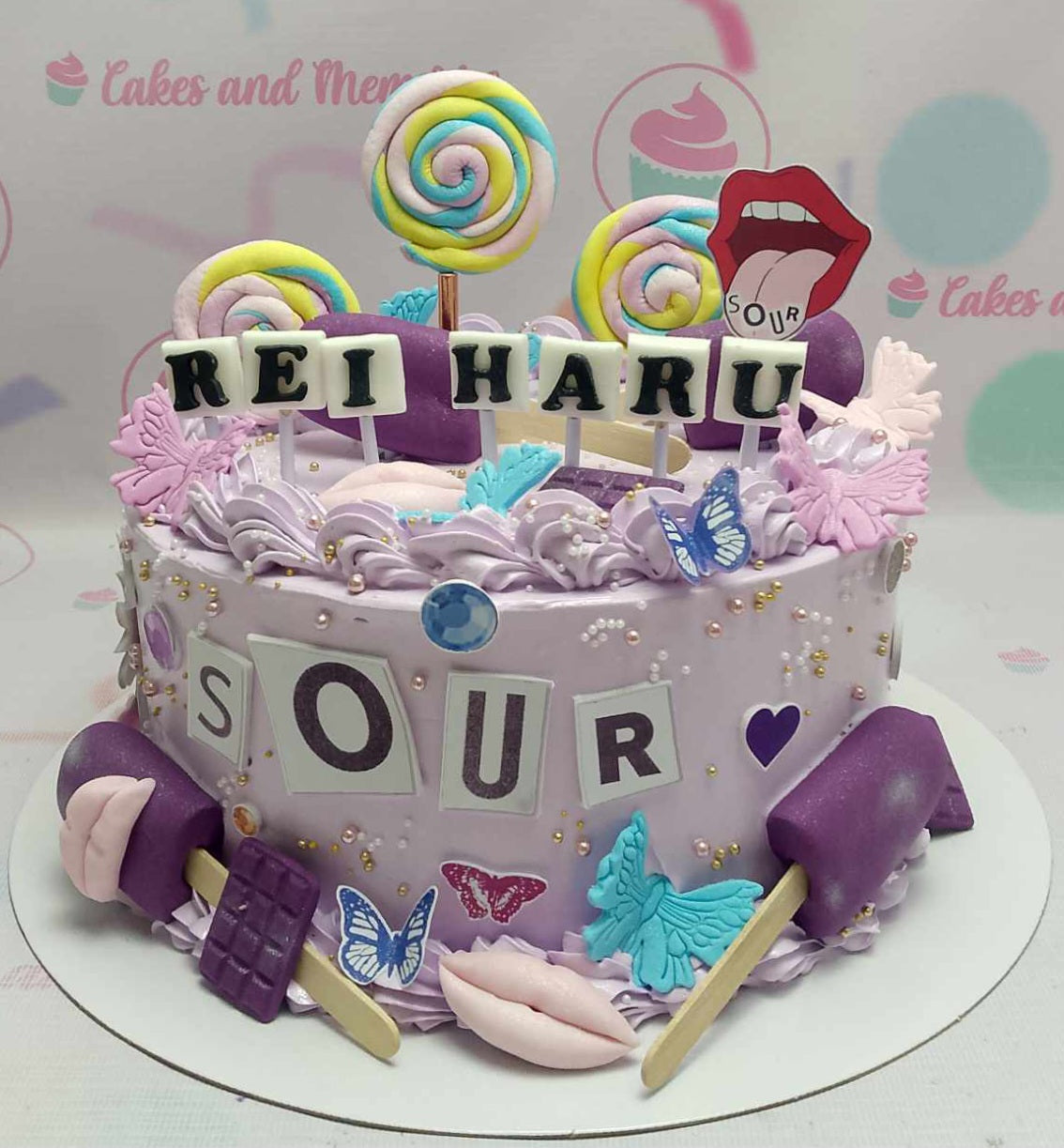 This custom Olivia Rodrigo-themed cake features a striking purple design adorned with printout toppers that include lollipops, butterflies, and a chocolate bar, accentuated by gold sprinkles and playful sour album elements. The cake captures the essence of pop music with its heart and mouth tongue accents, perfect for fans celebrating milestones like "Deja Vu" and "Driver's License."