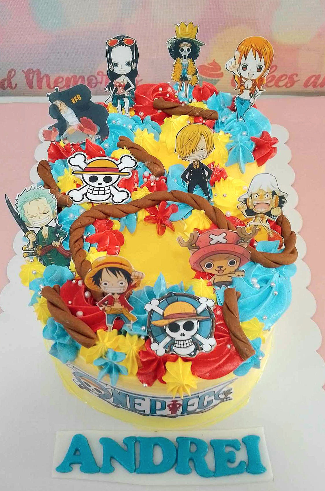 This custom One Piece cake features a rectangular design adorned with colorful printout toppers of Luffy, Zoro, Sanji, Chopper, and Nami, set against a backdrop of yellow, blue, and red. The cake showcases a playful skull and crossbones theme, complete with rope, stars, and pearls, perfect for Andrei's 6th birthday celebration.