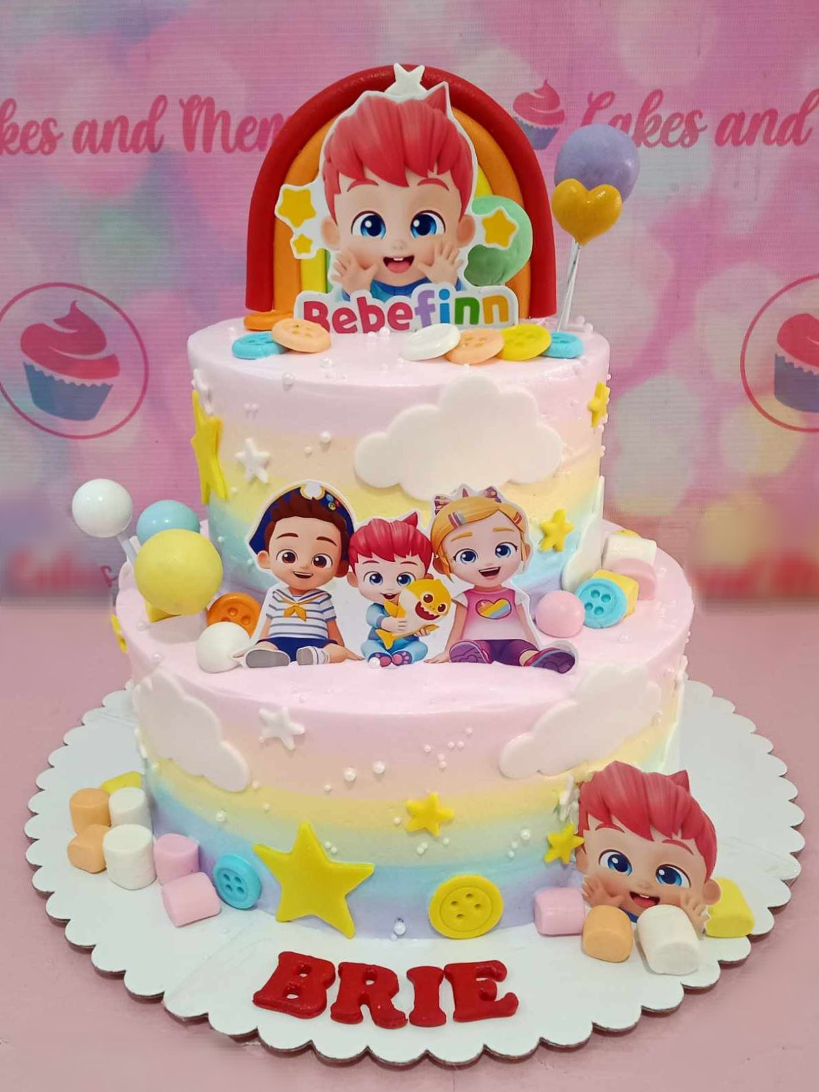 This delightful Bebefinn Cake features a two-tiered design adorned with colorful pastel hues and playful Bebefinn characters. Topped with an edible rainbow, stars, clouds, balloons, and buttons, it showcases charming printout toppers and miniature marshmallows for a fun celebration.