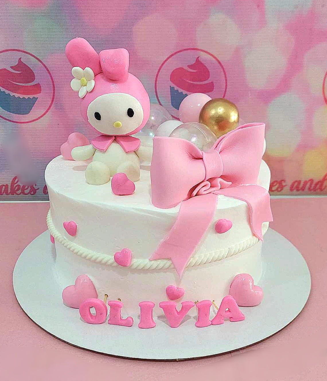 This charming My Melody Cake features a pink bow and ribbon, adorned with playful hearts and golden balls. Perfect for a girl's celebration, it showcases the beloved character surrounded by cute cartoon printout toppers.