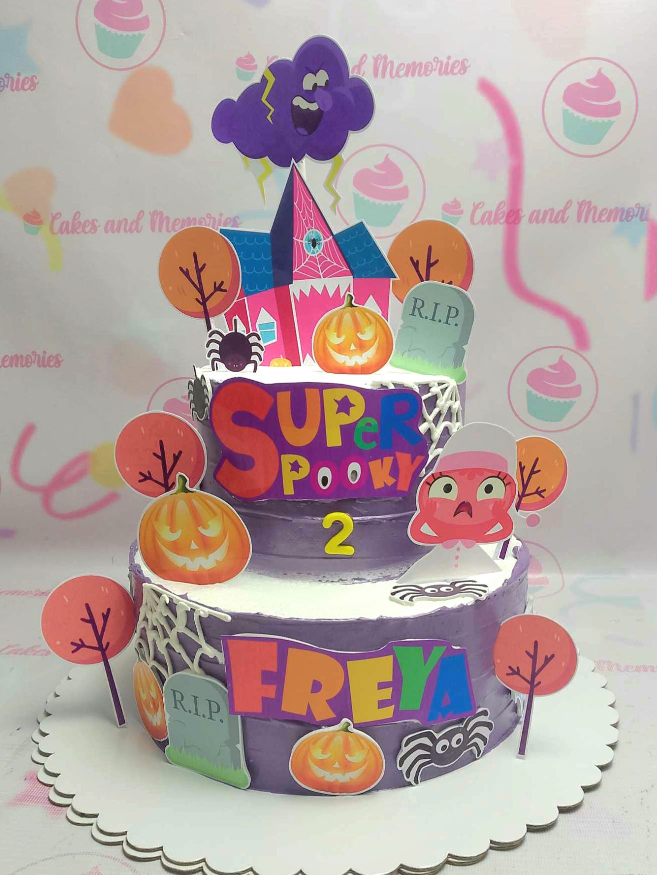 This two-tiered Super Simple Songs Cake features a playful design with purple hues and charming printout toppers, including a ghost, spider, haunted house, pumpkin, and tree, perfect for celebrating Freya's 2nd birthday. The graveyard scene adds a fun touch, making it an ideal centerpiece for toddlers who love baby songs and lullabies.