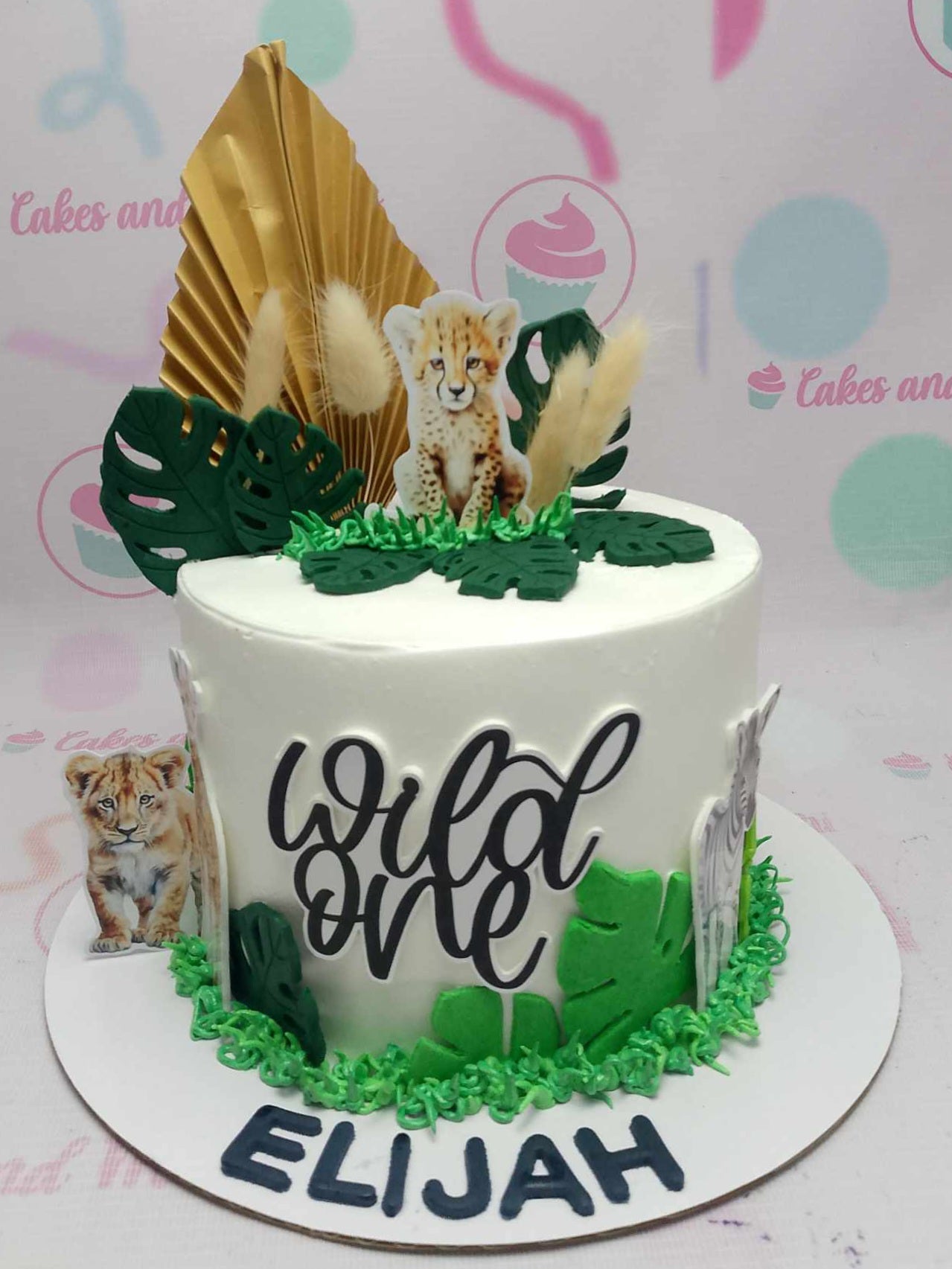 This vibrant 1-tier Safari Cake features a playful design with a white and green color palette, showcasing adorable animal printout toppers including a baby tiger, lion, zebra, and giraffe. Perfect for celebrating a "Wild One" 1st birthday, it is adorned with a gold fan for an extra touch of jungle charm.