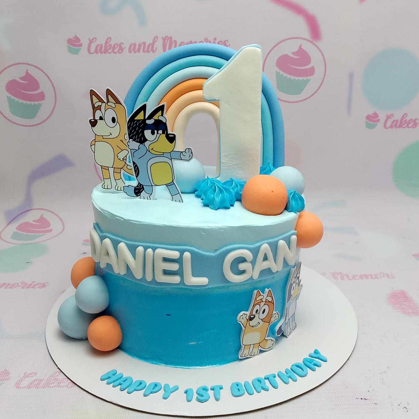 A vibrant Bluey Cake featuring various blue shades and a playful rainbow design, accented with blue and orange balls. This one-tier, one-layer birthday cake is adorned with printout toppers celebrating beloved toddler cartoons, perfect for any special occasion.