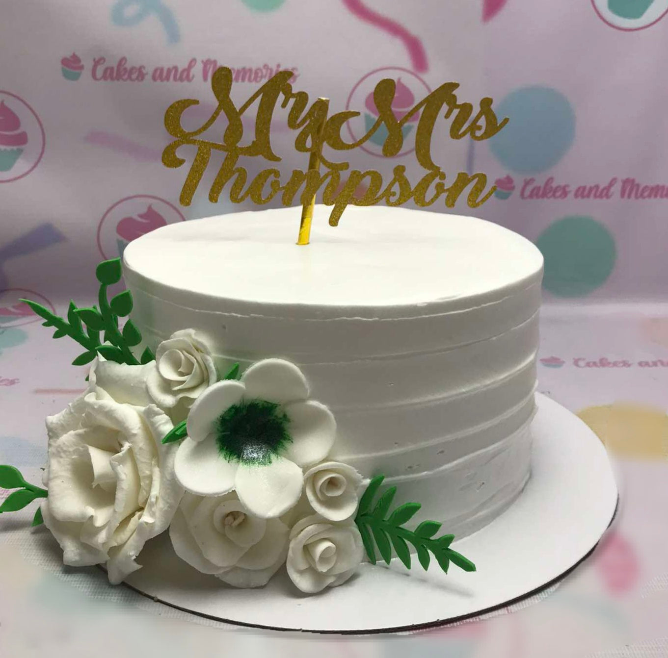 This elegant white wedding cake features a gold glitter cardstock topper and delicate white flowers, perfect for celebrating a 20th, 30th, 40th, 50th, or 60th anniversary. The single tier design makes it a lovely centerpiece for any wedding or birthday celebration.