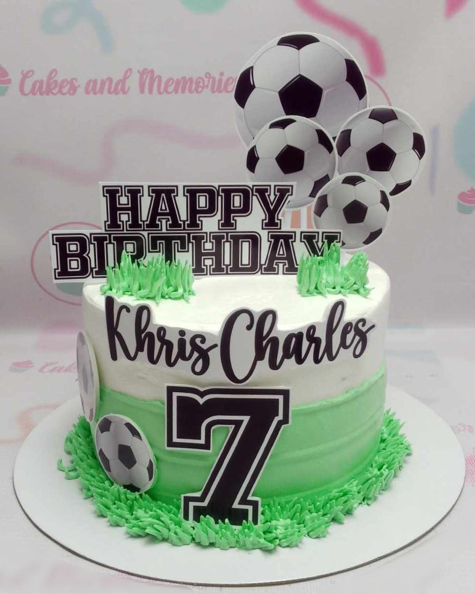 This custom Soccer Cake features a clean white base adorned with green grass accents and playful soccer ball decorations, perfect for celebrating Khris Charles' 7th birthday. The design includes printout toppers and highlights the number 7, making it an ideal centerpiece for any football-themed party.