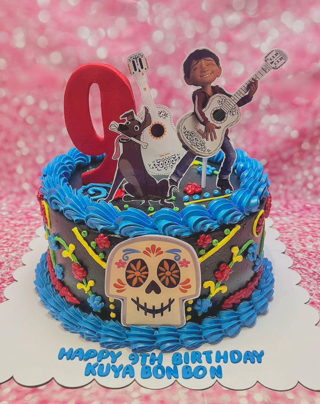 This custom Coco Cake features rich black icing accented with blue flowers, showcasing Miguel and Dante with a guitar and skull motifs. Topped with printout decorations, it celebrates a 9th birthday in honor of Kuya Bonbon, perfect for a Dia de Muertos theme.