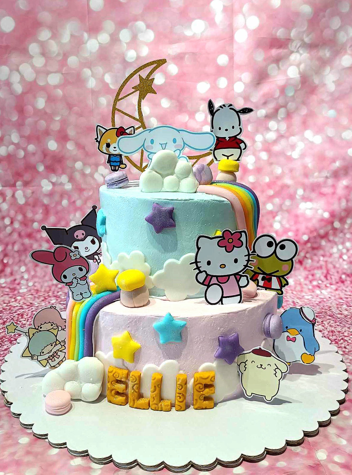 This charming Hello Kitty cake features light blue and purple colors across two tiers, adorned with Sanrio characters like Keroppi and My Melody, along with decorative stars, a rainbow, and a crescent moon. The delightful design is completed with clouds, mushrooms, and macarons, making it the perfect centerpiece for the birthday girl celebrating with Hello Kitty and friends.