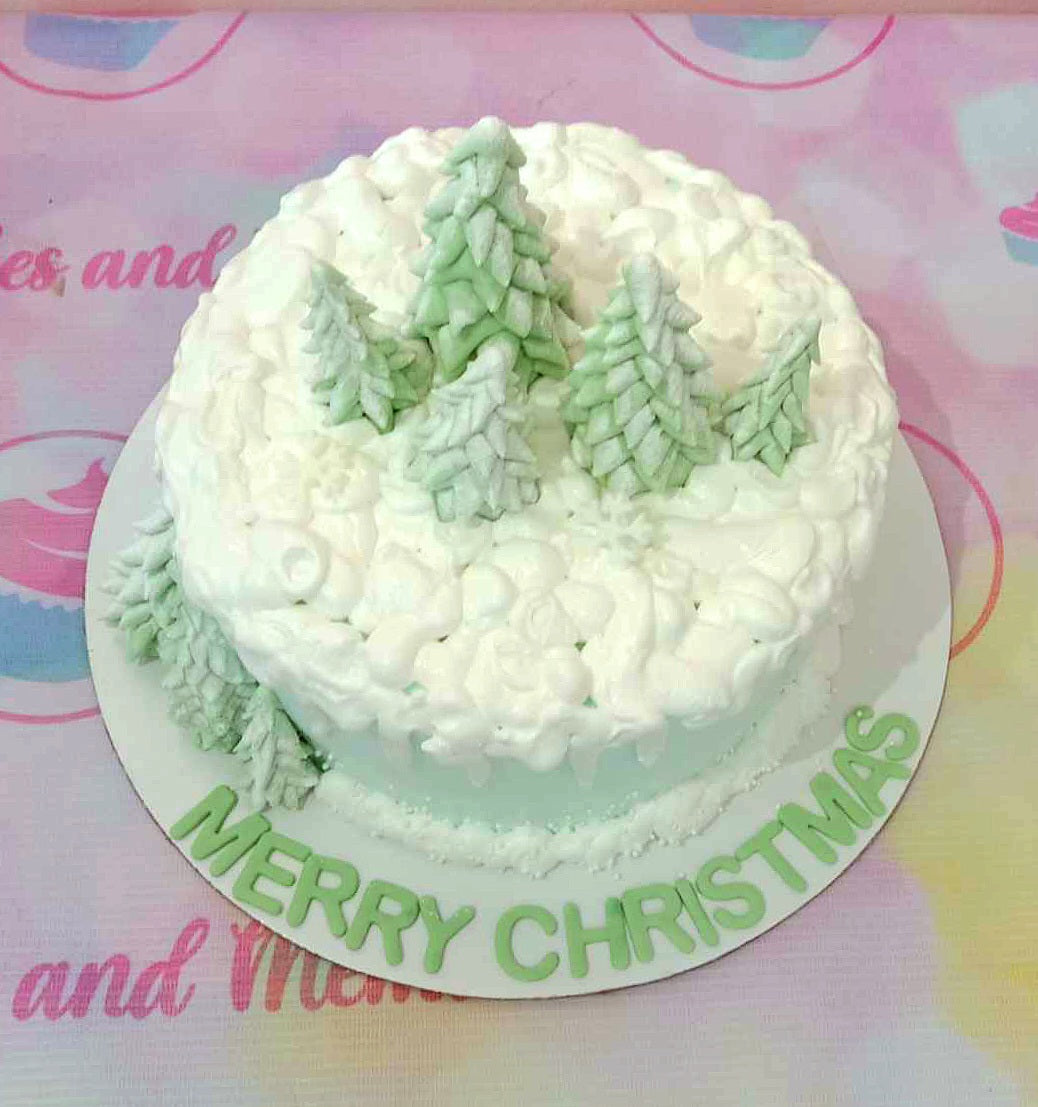 A beautifully crafted light green Christmas cake featuring a stunning Christmas tree design set in a winter wonderland backdrop of snow and pine trees. Perfect for your Christmas party, this one-tier cake captures the festive spirit with a "Merry Christmas" message.