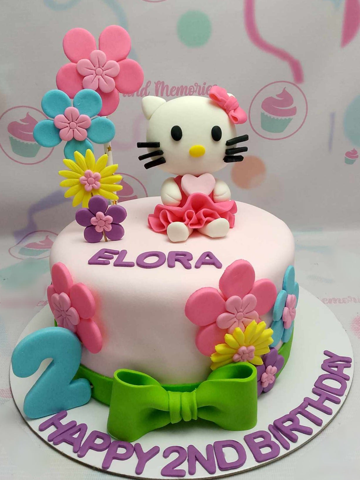 This charming Hello Kitty Cake features a delightful pink and white design adorned with a light green ribbon, flowers, and a bow, perfect for celebrating Elora's 2nd birthday. The edible topper showcases Hello Kitty and the number 2, making it an ideal centerpiece for a baby girl's special day.