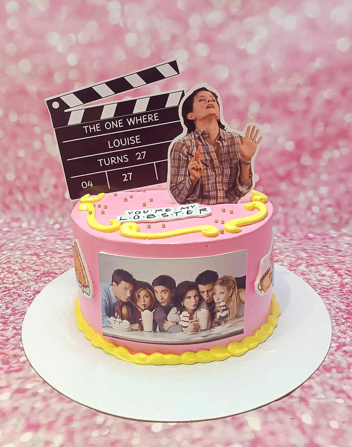 This Friends Cake features delightful pink icing, adorned with a clapper movie slate and the name "Monica" with the number 7, perfect for a 27th birthday girl. The single-tier design showcases custom printout toppers inspired by the beloved Friends TV show series.