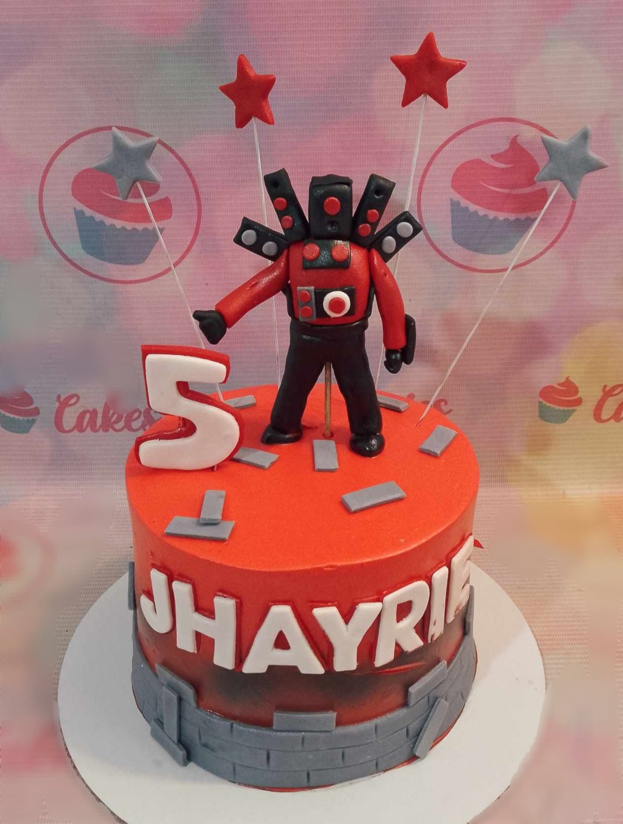 Celebrate a 5th birthday with this vibrant Skibidi Cake featuring a striking red design adorned with grey brick patterns. Topped with playful printout toppers of Titan Speakerman and stars, this whimsical one-tier masterpiece is perfect for Gen Z and Gen Alpha video game enthusiasts.