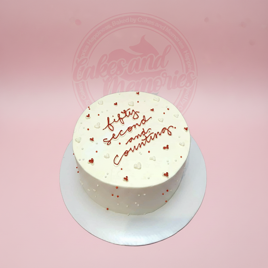Round anniversary cake with 'fifty second and counting' text, red and white piped hearts, and sprinkles on white buttercream.
