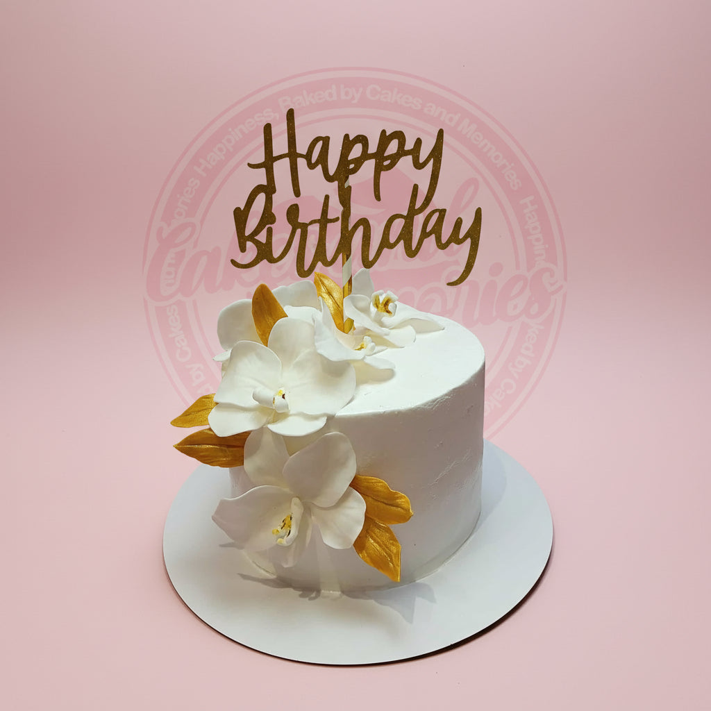 One-tier birthday cake with elegant white sugar orchids, gold leaves, and 'Happy Birthday' topper on a pink background.