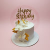 One-tier birthday cake with elegant white sugar orchids, gold leaves, and 'Happy Birthday' topper on a pink background.