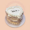 White gender reveal cake with 'it's a ...' text and elegant buttercream piping, perfect for baby announcements.