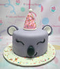 A charming Koala Cake featuring a gray koala bear adorned with a pink party hat and surrounded by colorful candy sprinkles. This 1-layer, 1-tier masterpiece is crafted with fondant, perfect for any celebration.