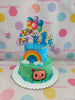 Two-tier Cocomelon 1st birthday cake with blue and green buttercream, 'Happy Birthday Caleb Jiro!' topper, and character printouts.